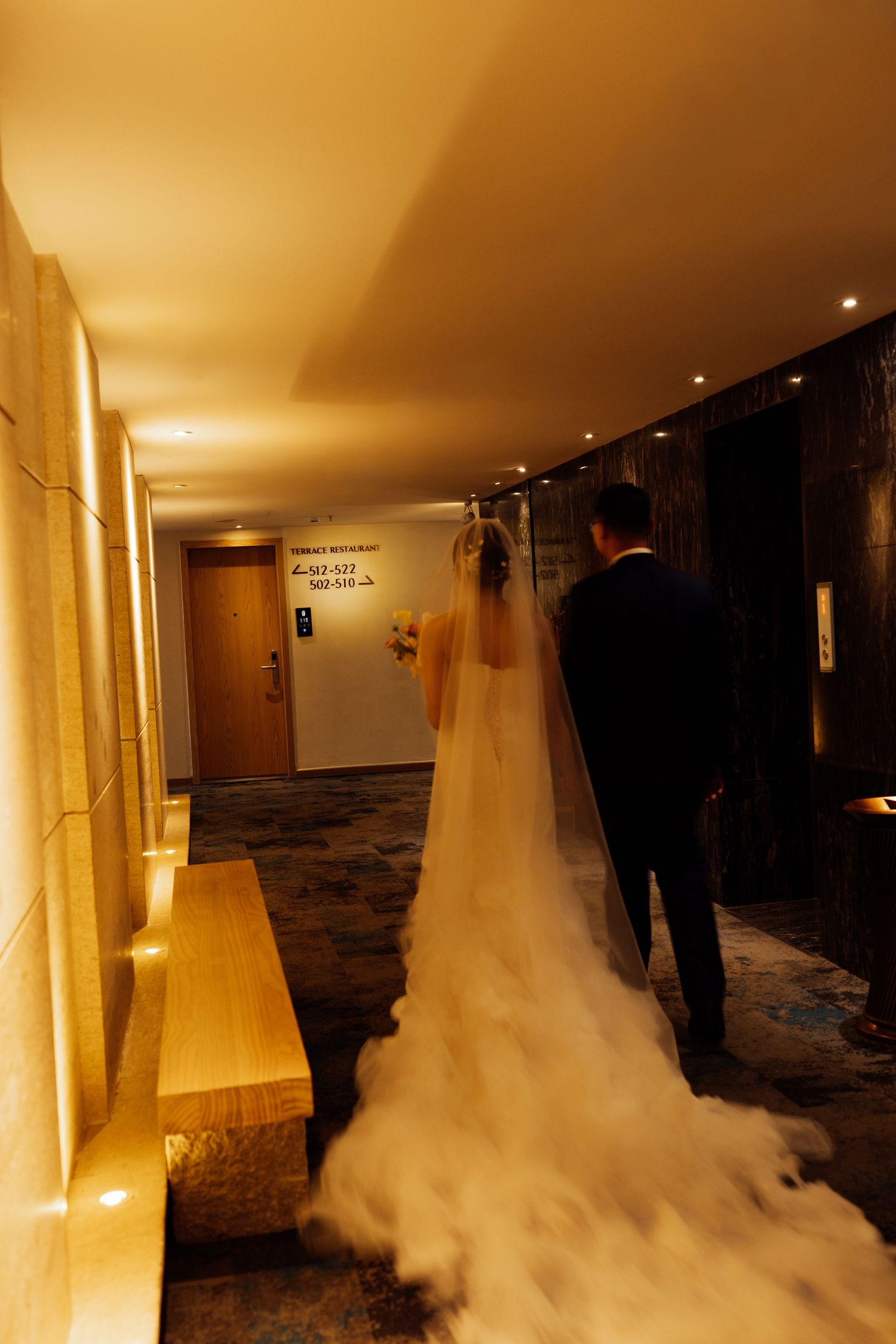 Bride and groom walking down a warmly lit hotel corridor, her white gown and veil trailing elegantly behind them.