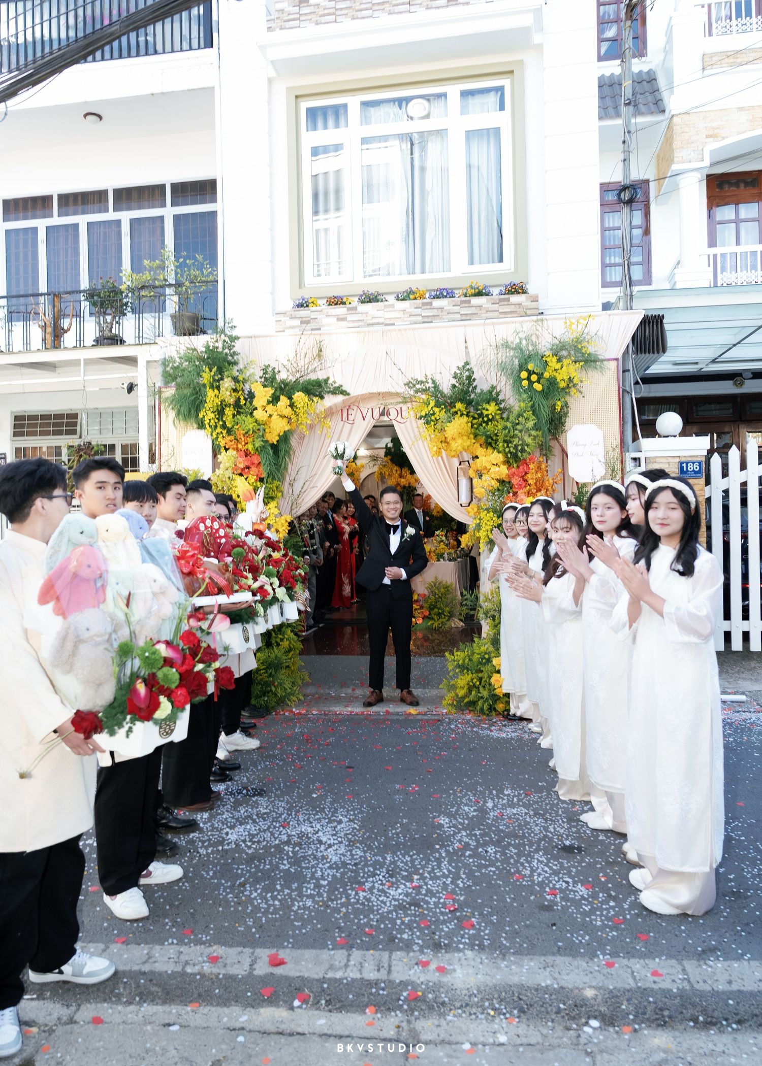 Groom walks through floral arch at Vietnamese wedding ceremony while guests in white ao dai clap and celebrate outdoors.