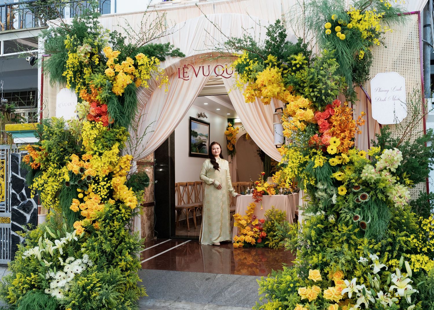 Woman in ao dai stands at floral archway entrance adorned with yellow and white blooms at Hieu Vu Quan restaurant.