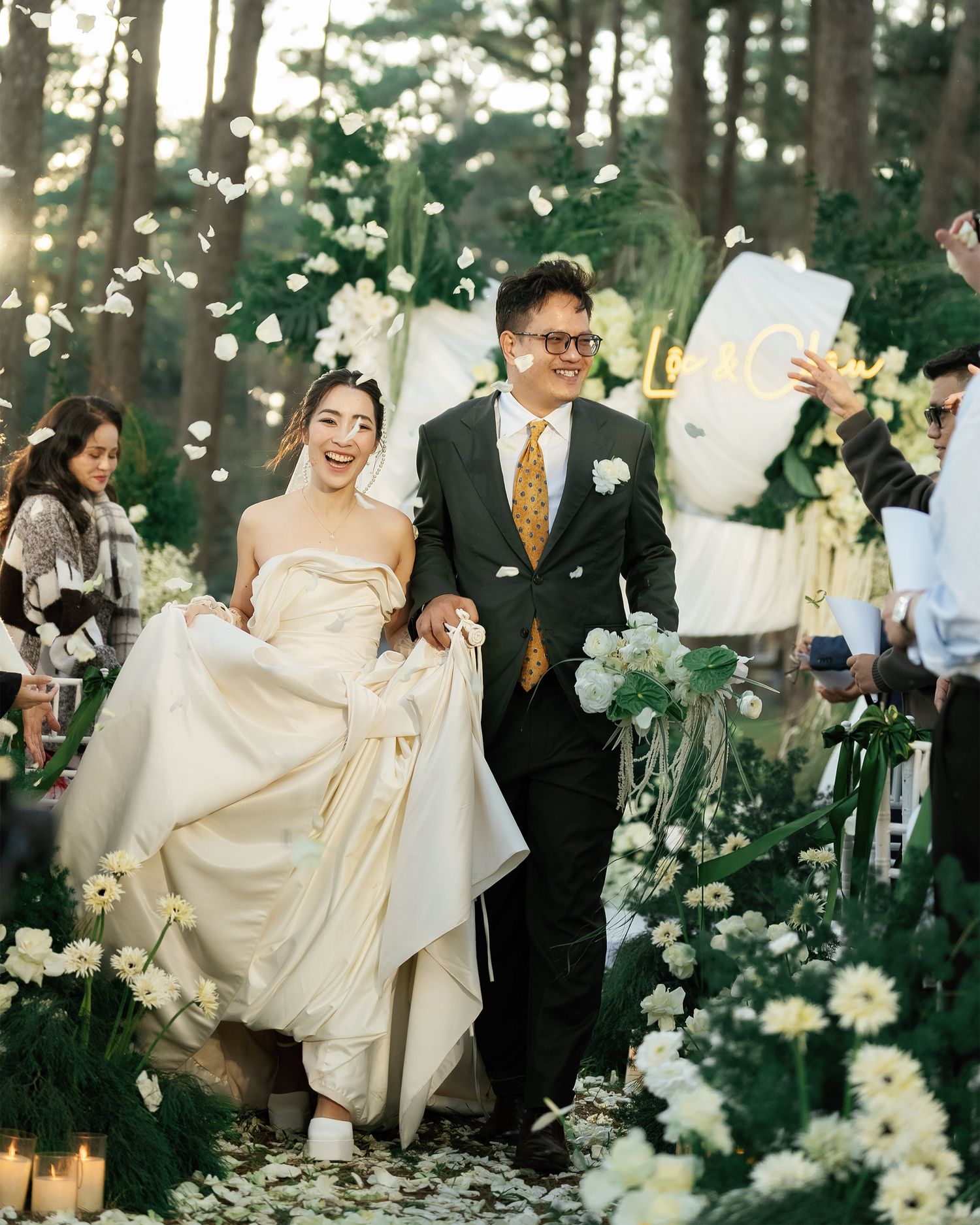 Happy couple walks down outdoor wedding aisle lined with white flowers and greenery in a forest setting.