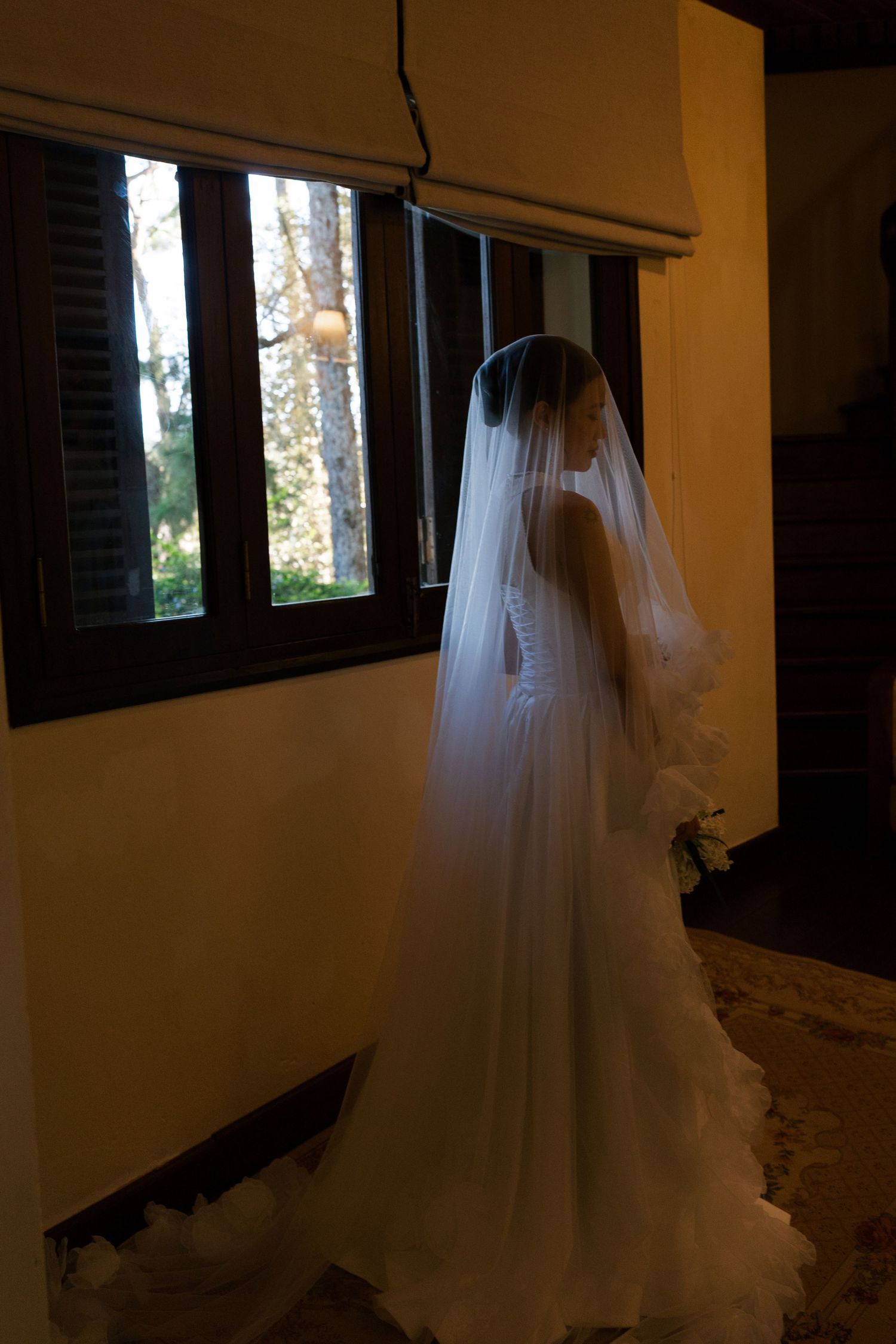 Silhouette of bride in wedding gown and veil standing by a window in soft natural light.