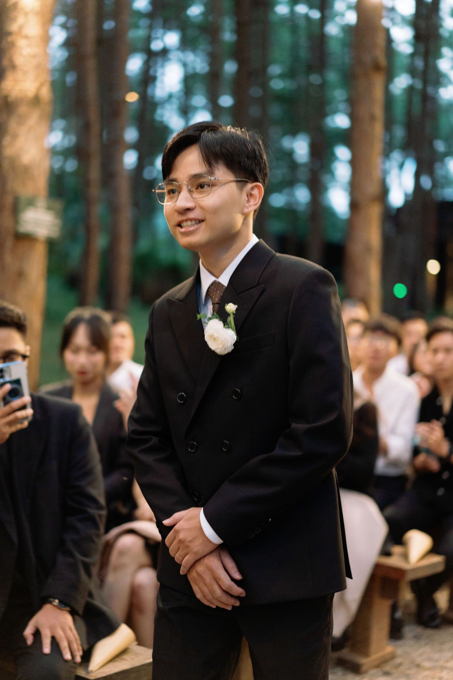 Groom in black suit with white boutonniere smiling at outdoor forest wedding ceremony.