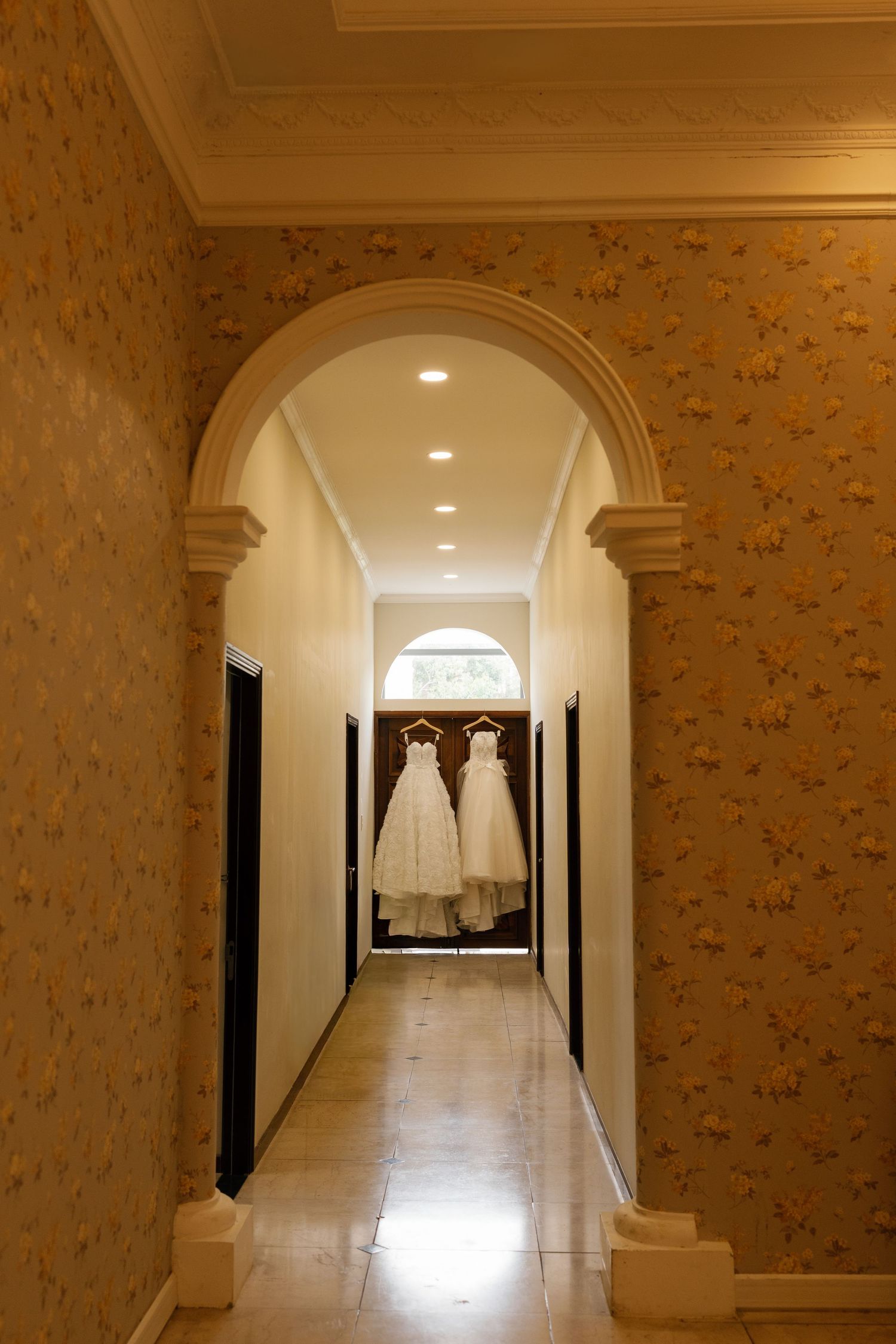 Elegant hallway with ornate arch, gold floral wallpaper, and two white wedding dresses hanging near a window.