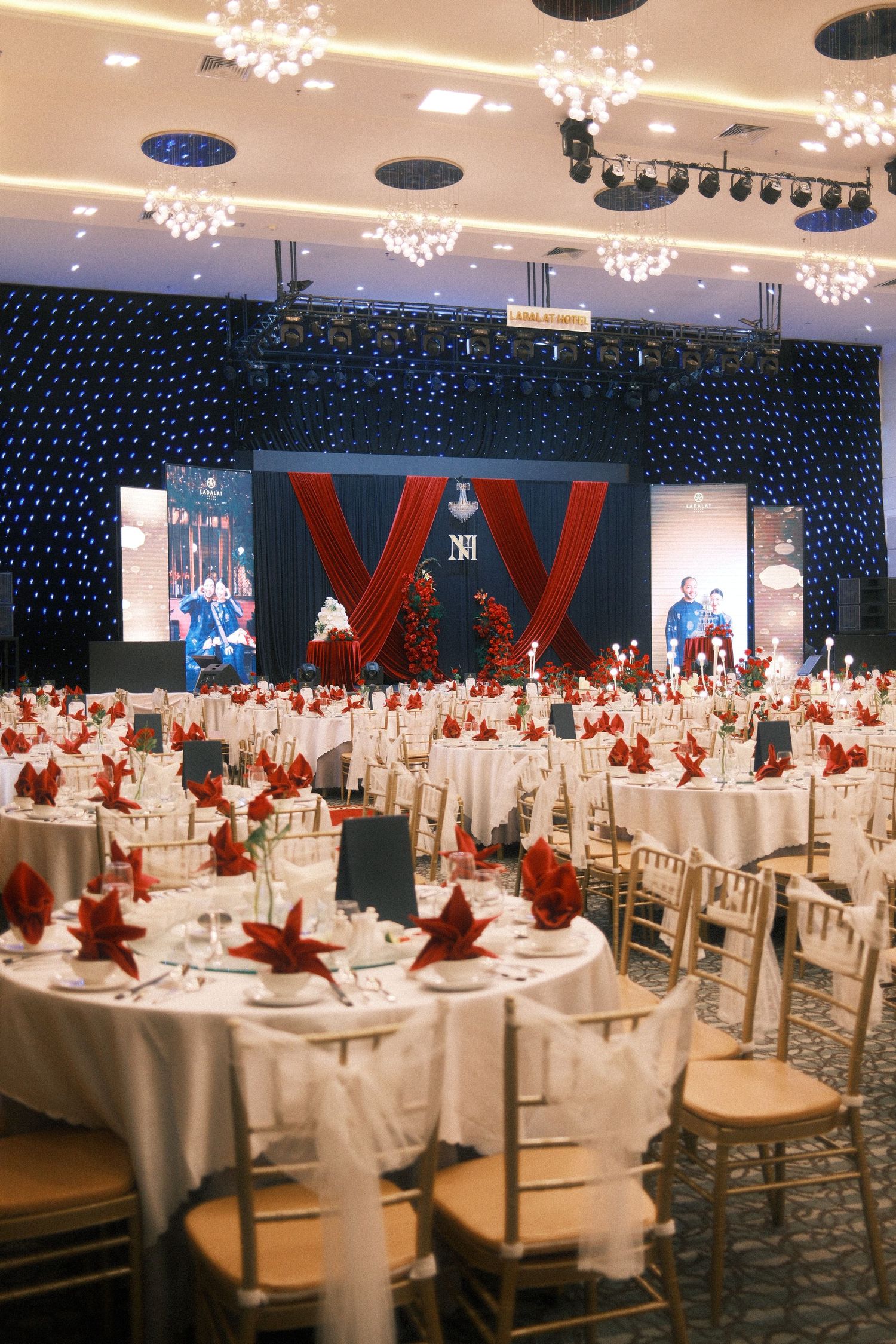 Elegant banquet hall with round tables, red napkins, gold chairs, and a decorated stage with red drapes.