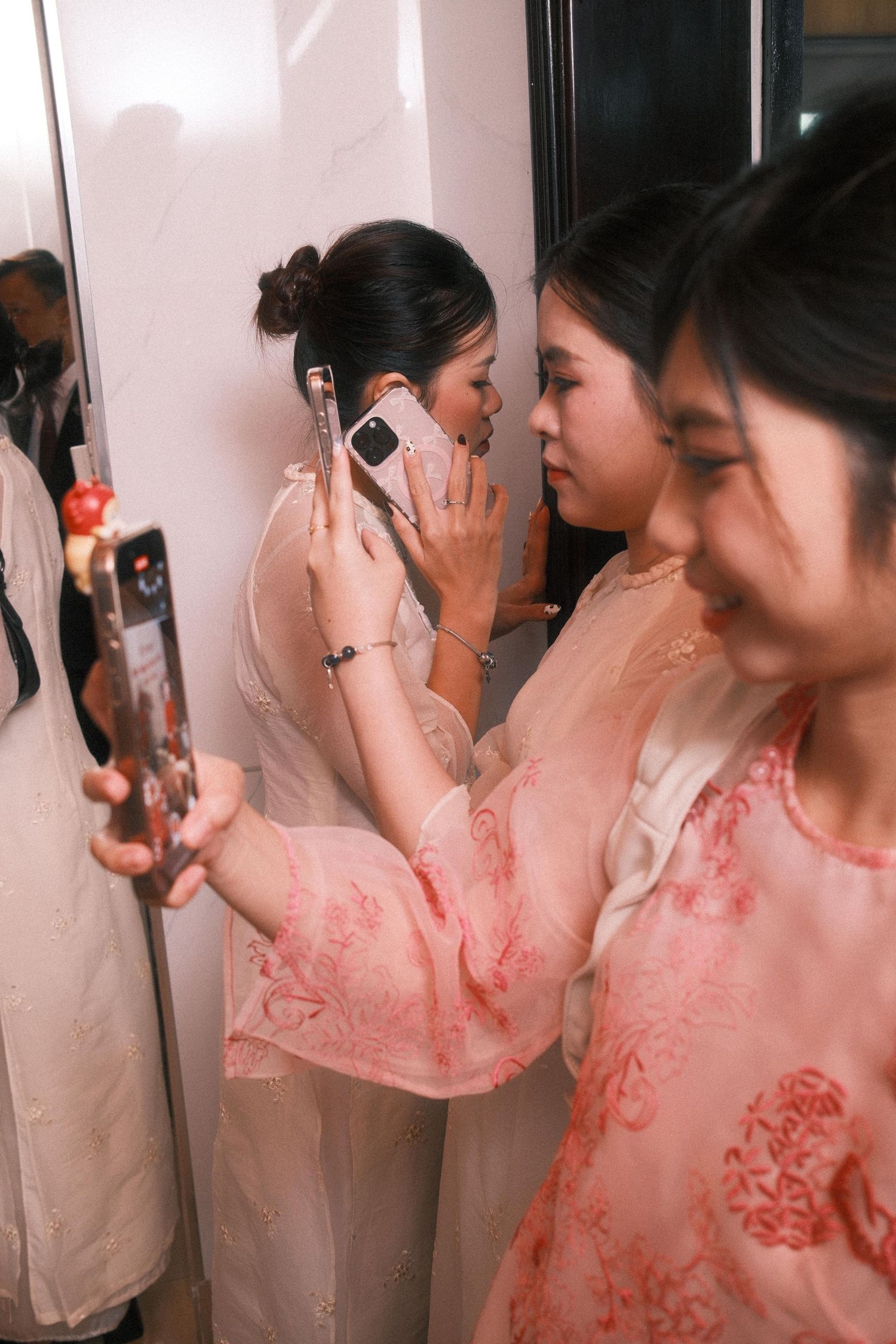 Two Asian women in pink floral robes taking selfies with smartphones near a mirror at what appears to be a wedding.
