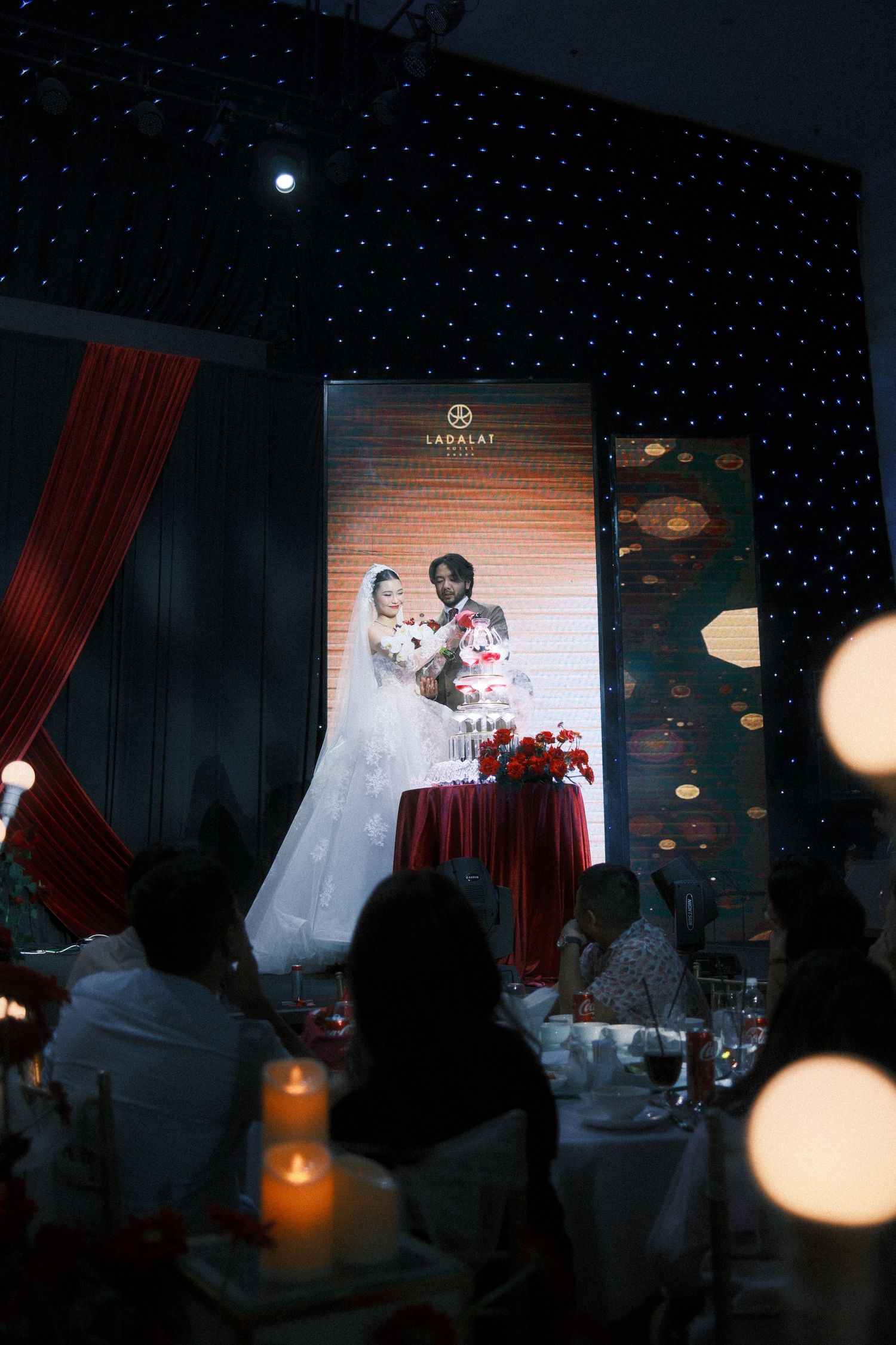 Bride and groom cutting wedding cake at elegant reception with candlelit tables and starry backdrop.