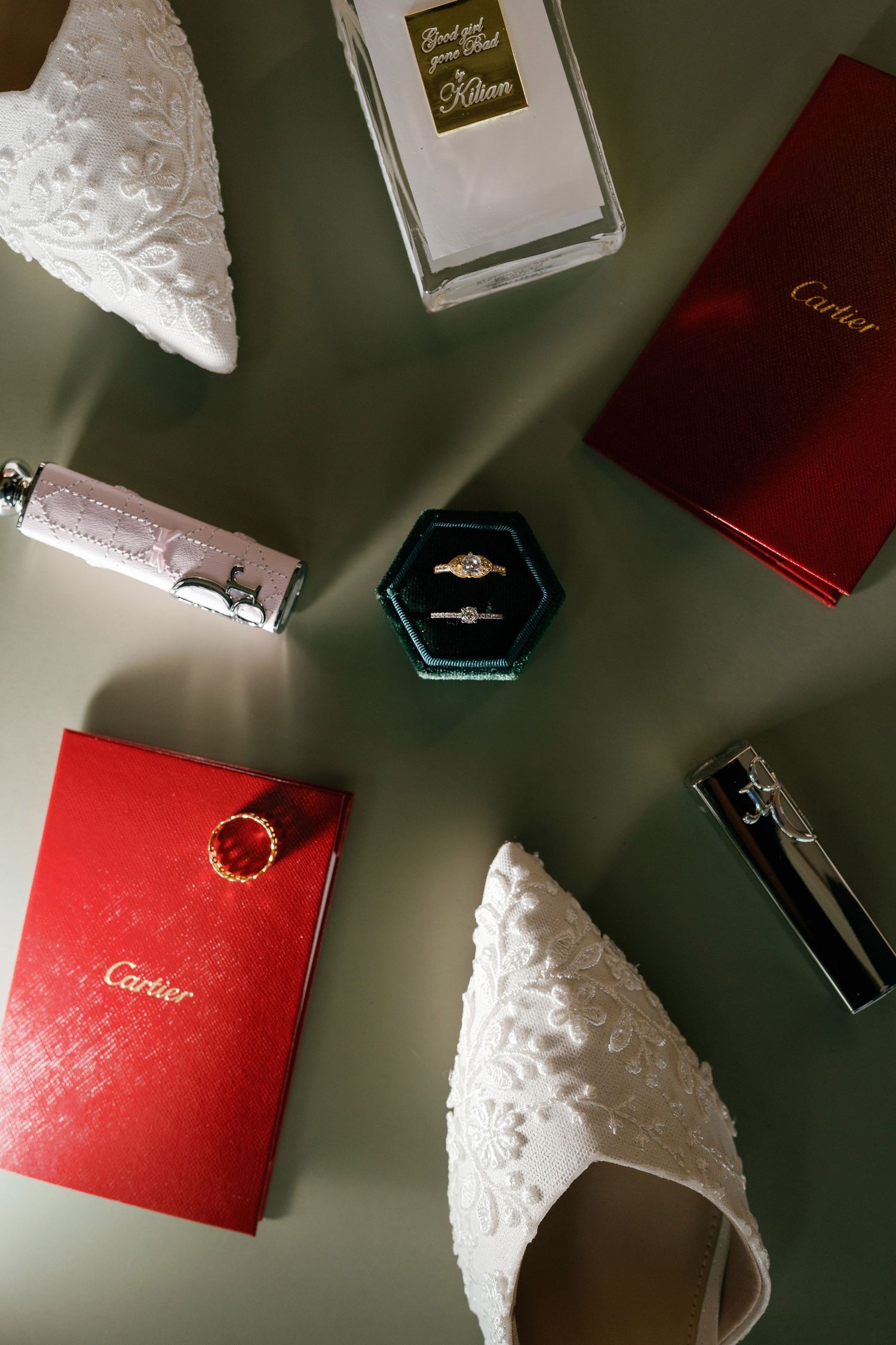 Bridal flat lay featuring lace heels, Cartier boxes, wedding rings in teal box, Killian perfume on sage green background.