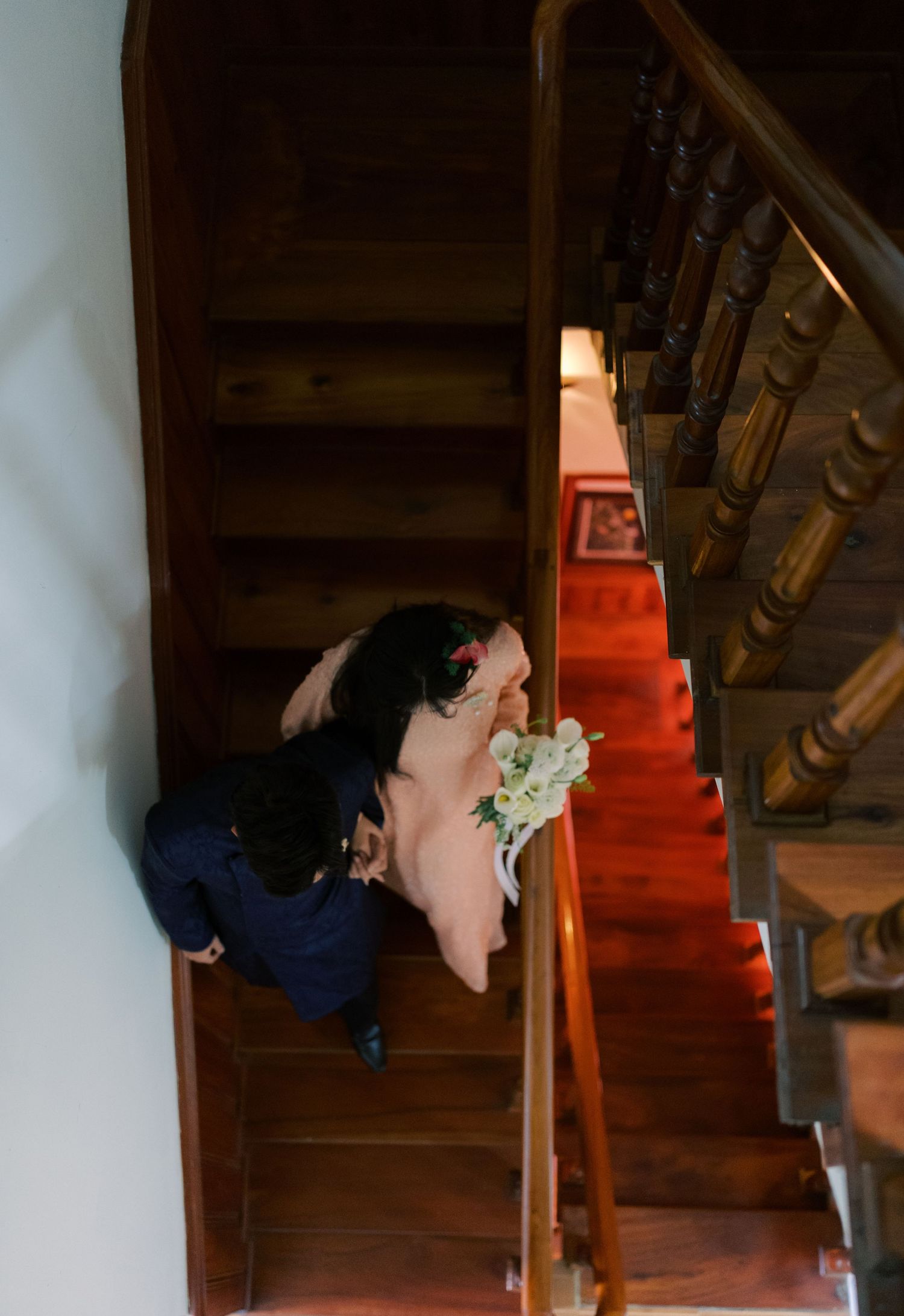 Aerial view of bride and groom descending wooden staircase with red carpet, bride holding white bouquet.