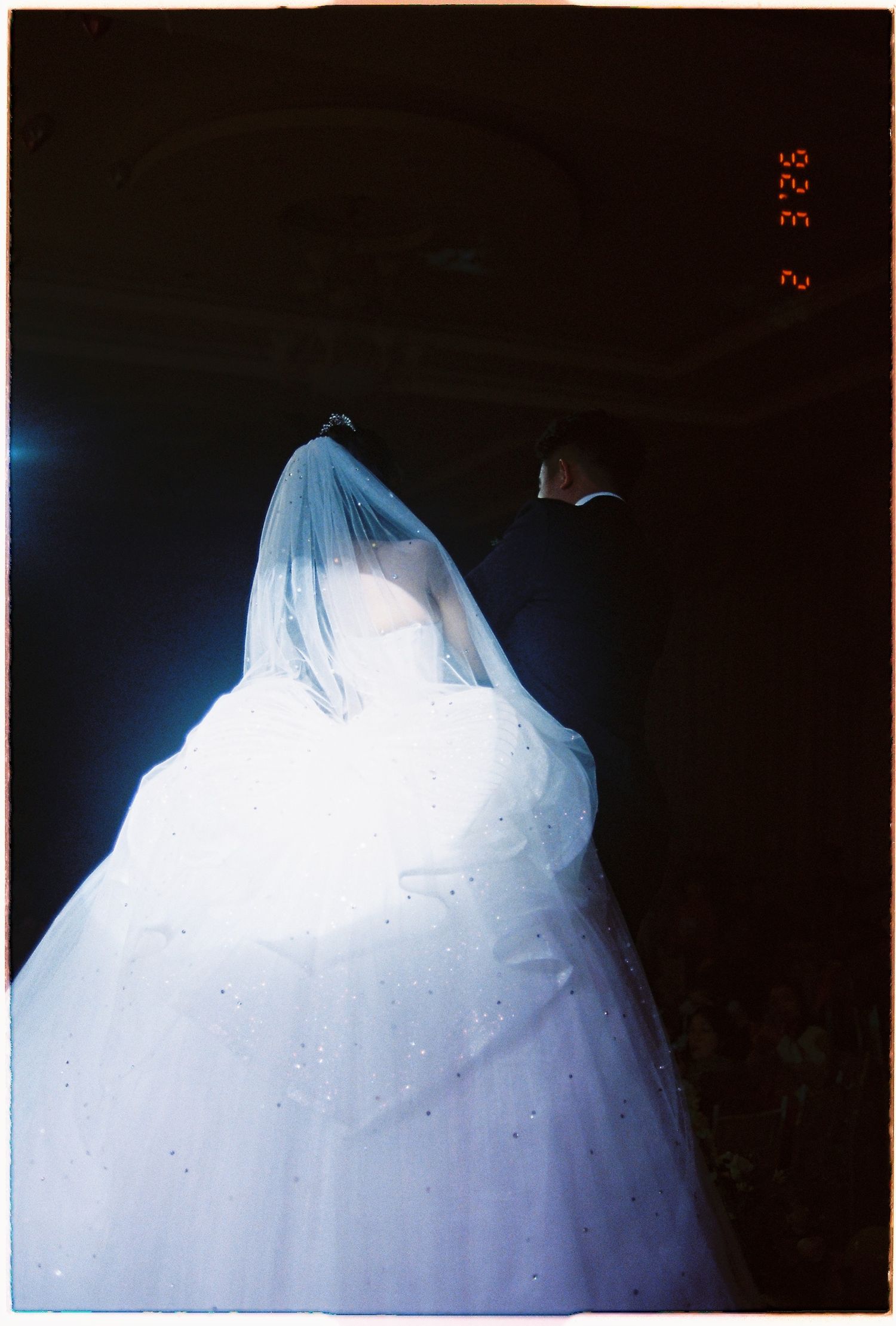 Artistic film photo of Vietnamese bride in white ballgown and veil with groom in dark background.