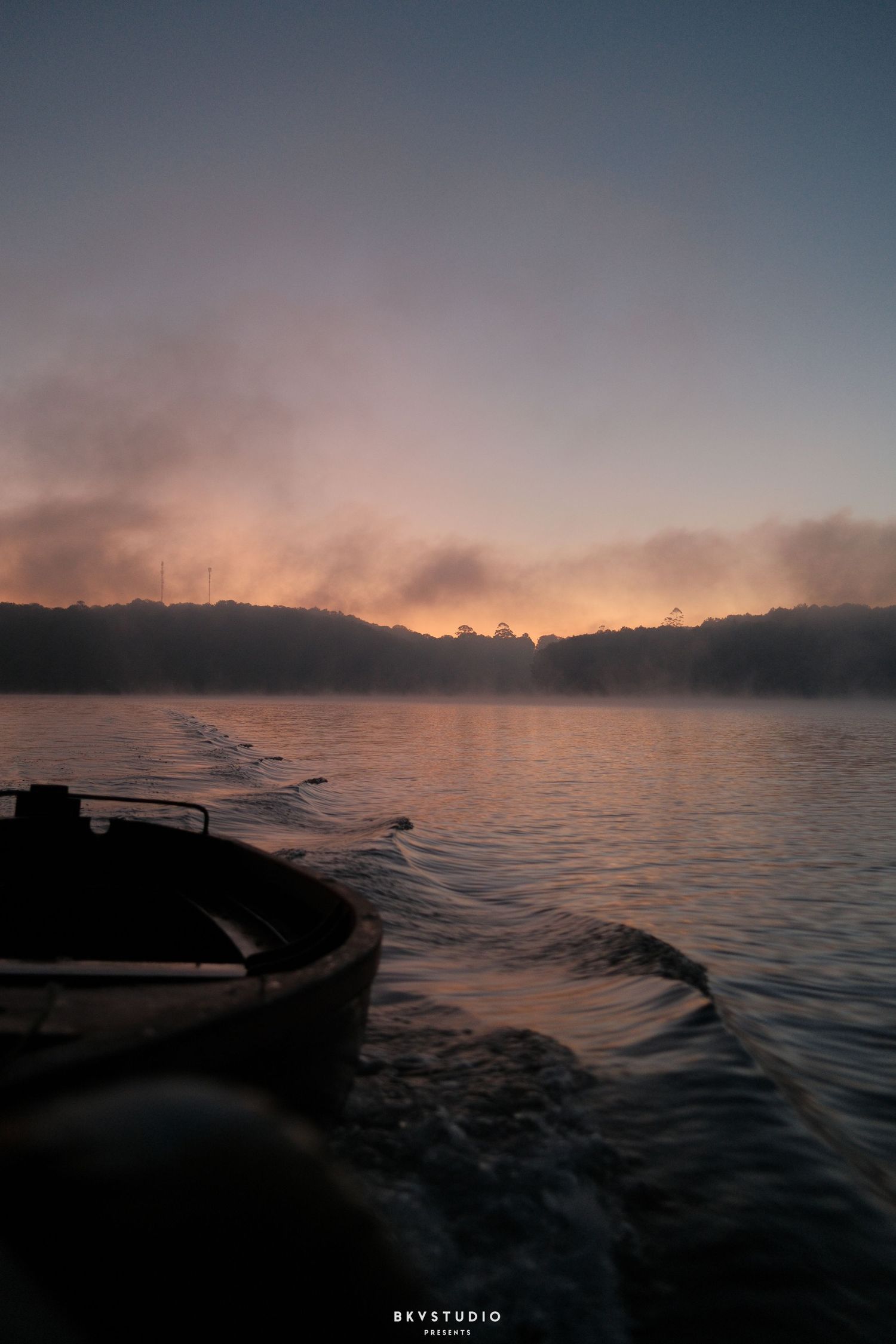 A misty sunrise over a tranquil lake with a wooden boat in the foreground and forested hills in the background.