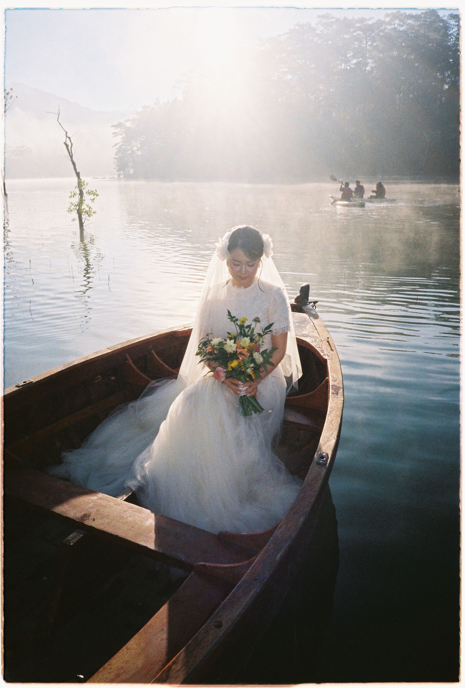Bride in white gown and veil sits alone in wooden boat on misty lake holding floral bouquet.