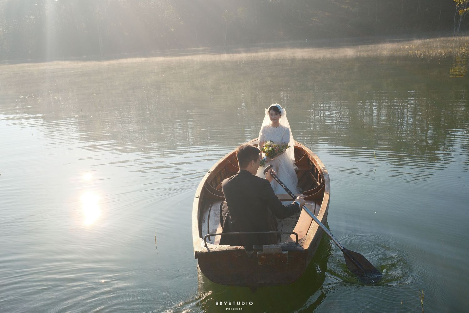 Bride and groom rowing a wooden boat on a calm misty lake surrounded by trees.