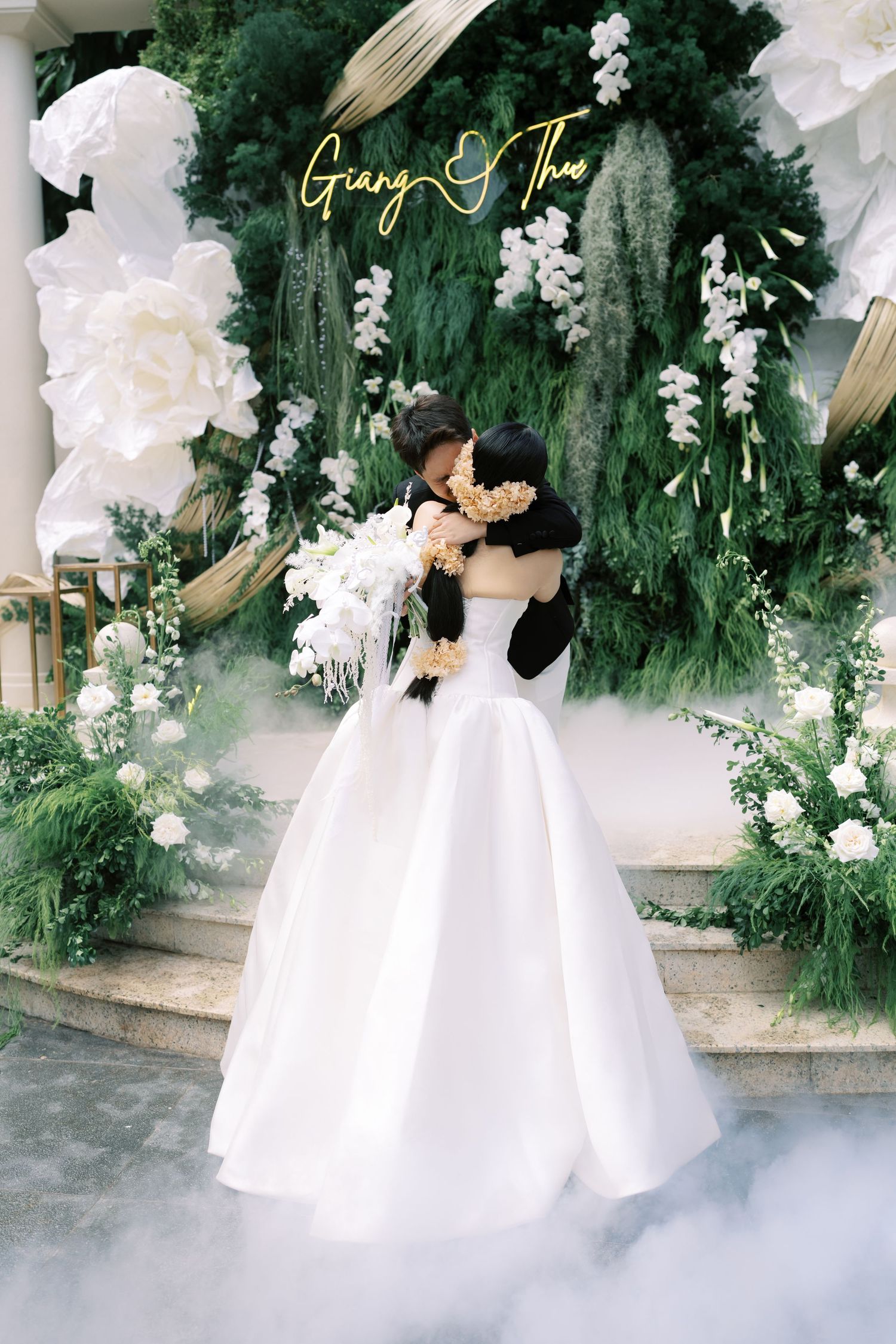 Bride and groom sharing romantic kiss under lush green floral backdrop at Giang and Thao wedding.