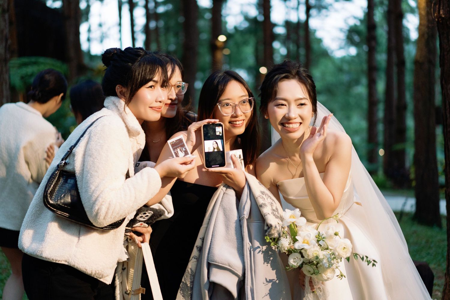 Bride in white gown poses with smiling guests viewing photos on phone at outdoor forest wedding.
