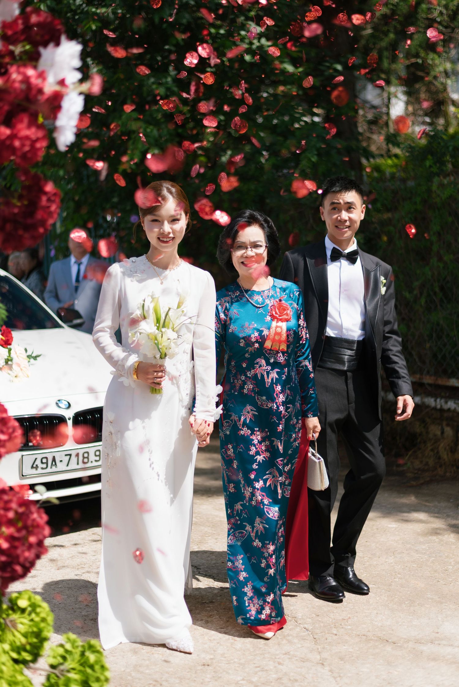 A bride in white ao dai holds flowers beside a woman in teal floral dress and a groom in tuxedo at a Vietnamese wedding.