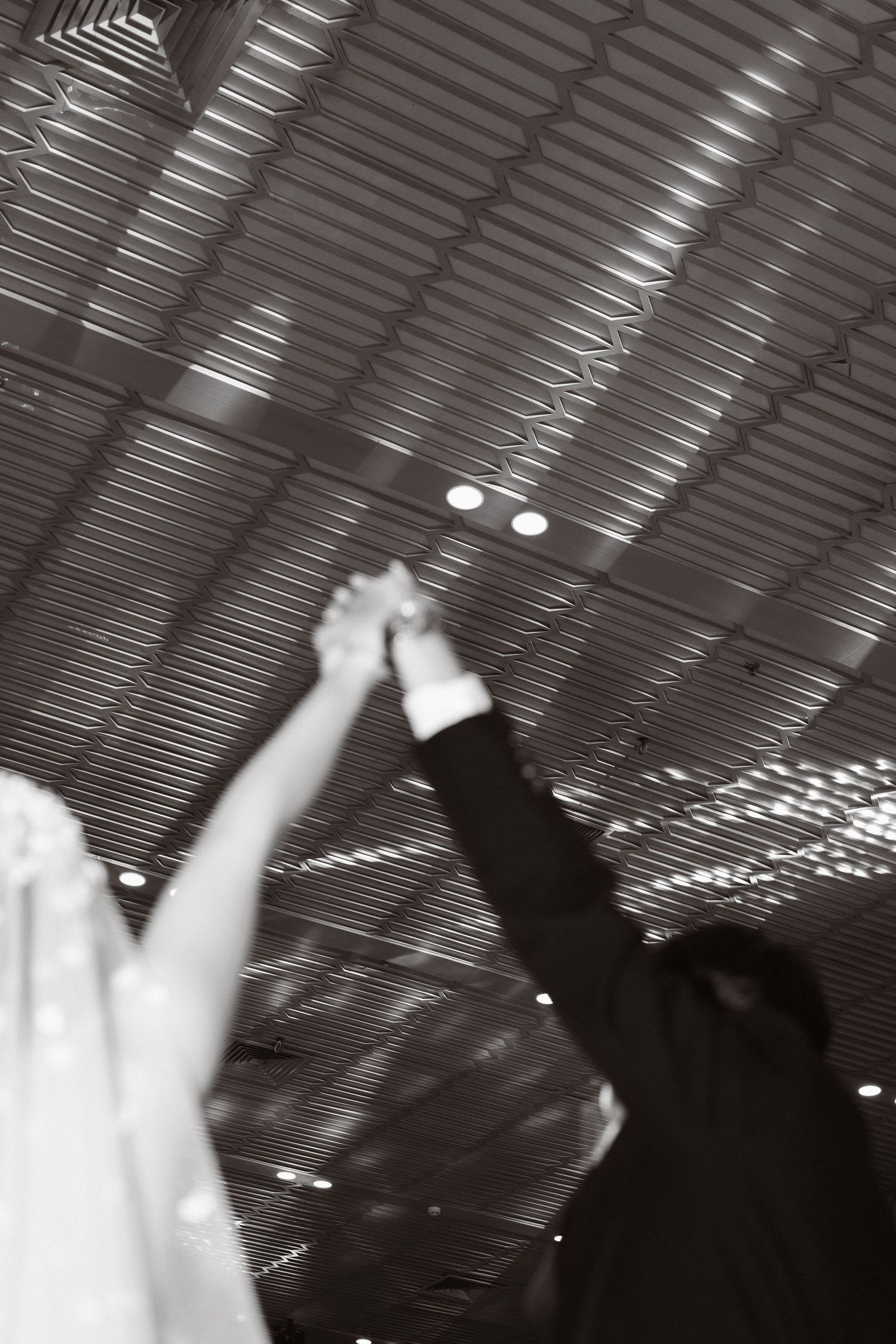 Bride and groom raising joined hands in celebration beneath a modern geometric ceiling at their wedding reception.