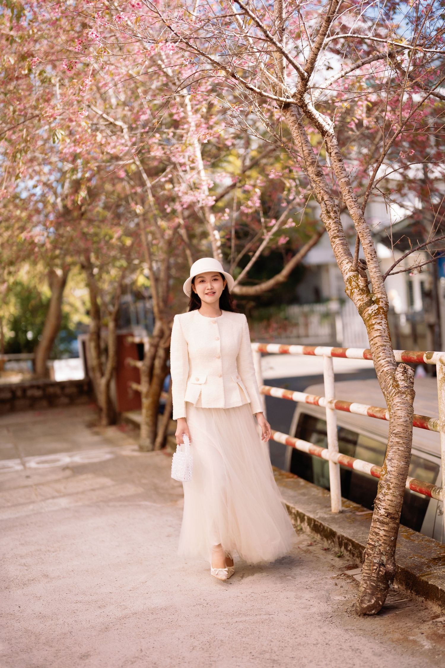 Woman in white peplum jacket and tulle skirt poses under blooming cherry blossom trees on a scenic walkway.