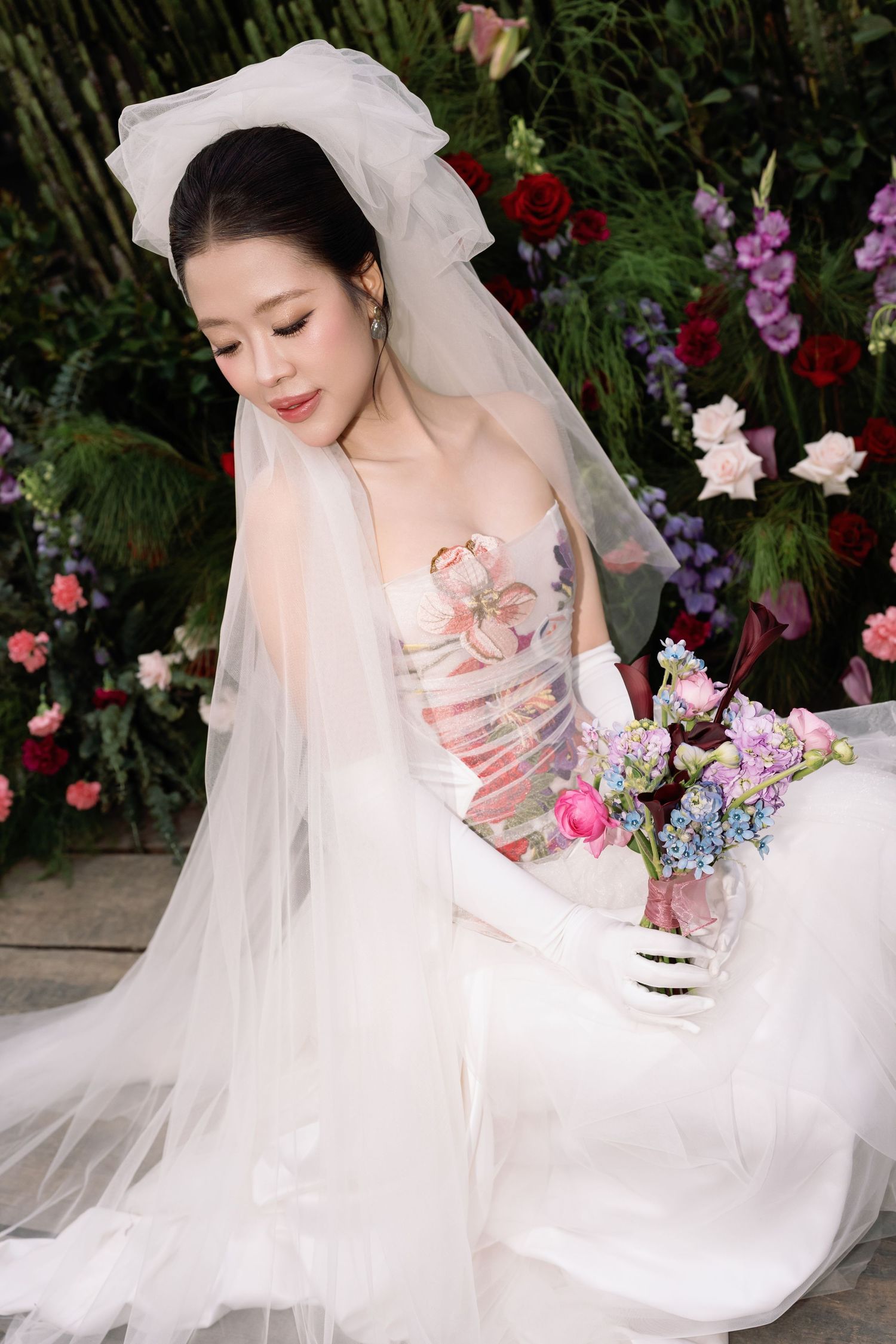 Elegant bride in floral strapless gown with white veil and gloves holding colorful bouquet among flowers.