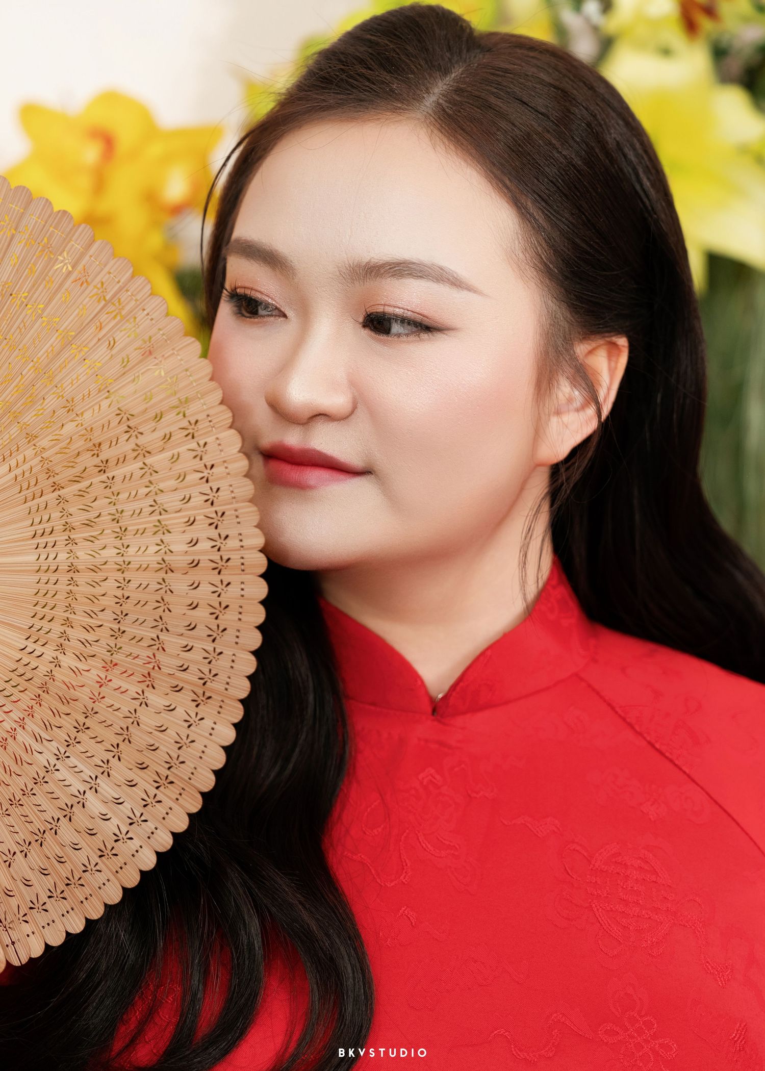 Young woman in red áo dài holding a woven fan, with elegant makeup and long black hair, posed before yellow flowers.