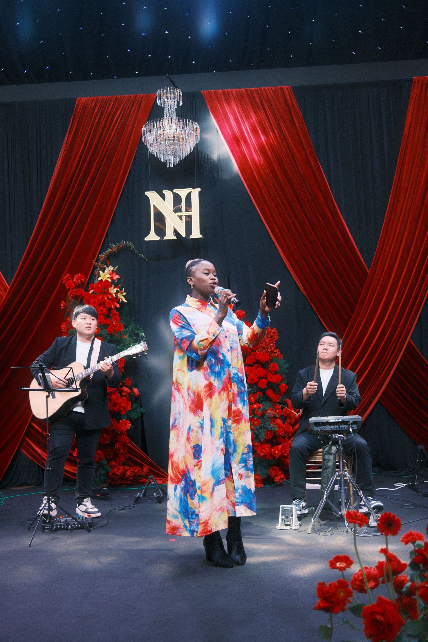 Singer in floral dress performs on stage with guitarist and keyboardist amid red curtains and roses.