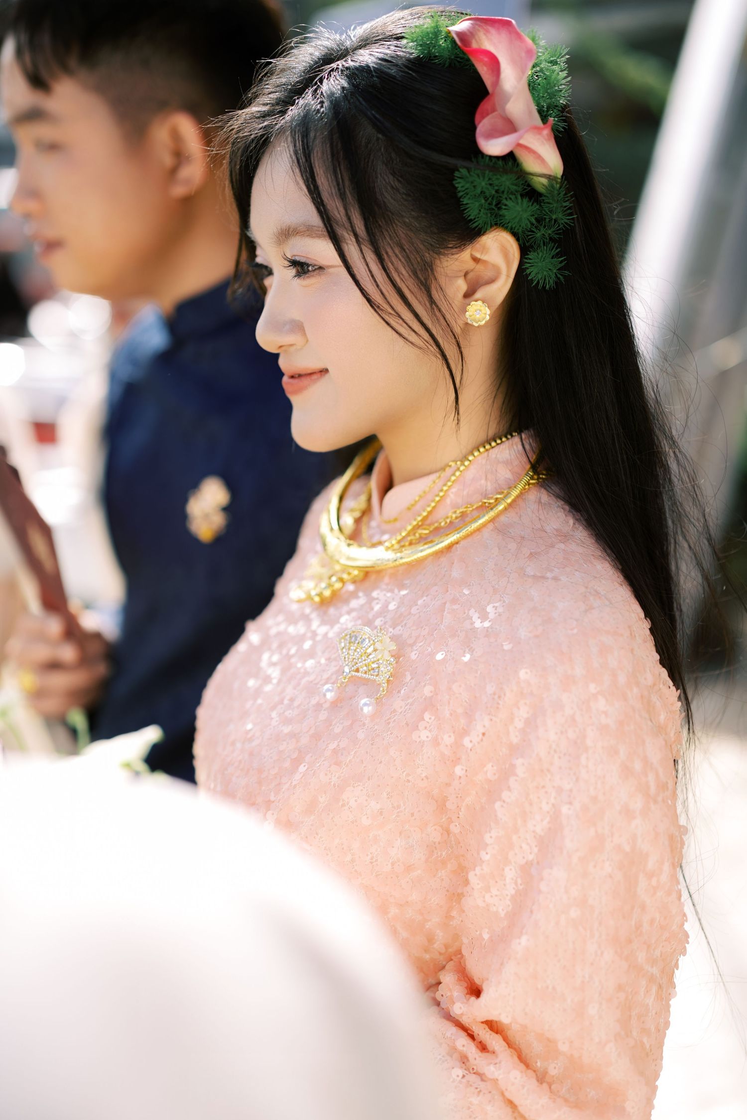 Vietnamese bride in pink ao dai with gold jewelry and pink flower hair accessory at wedding ceremony.