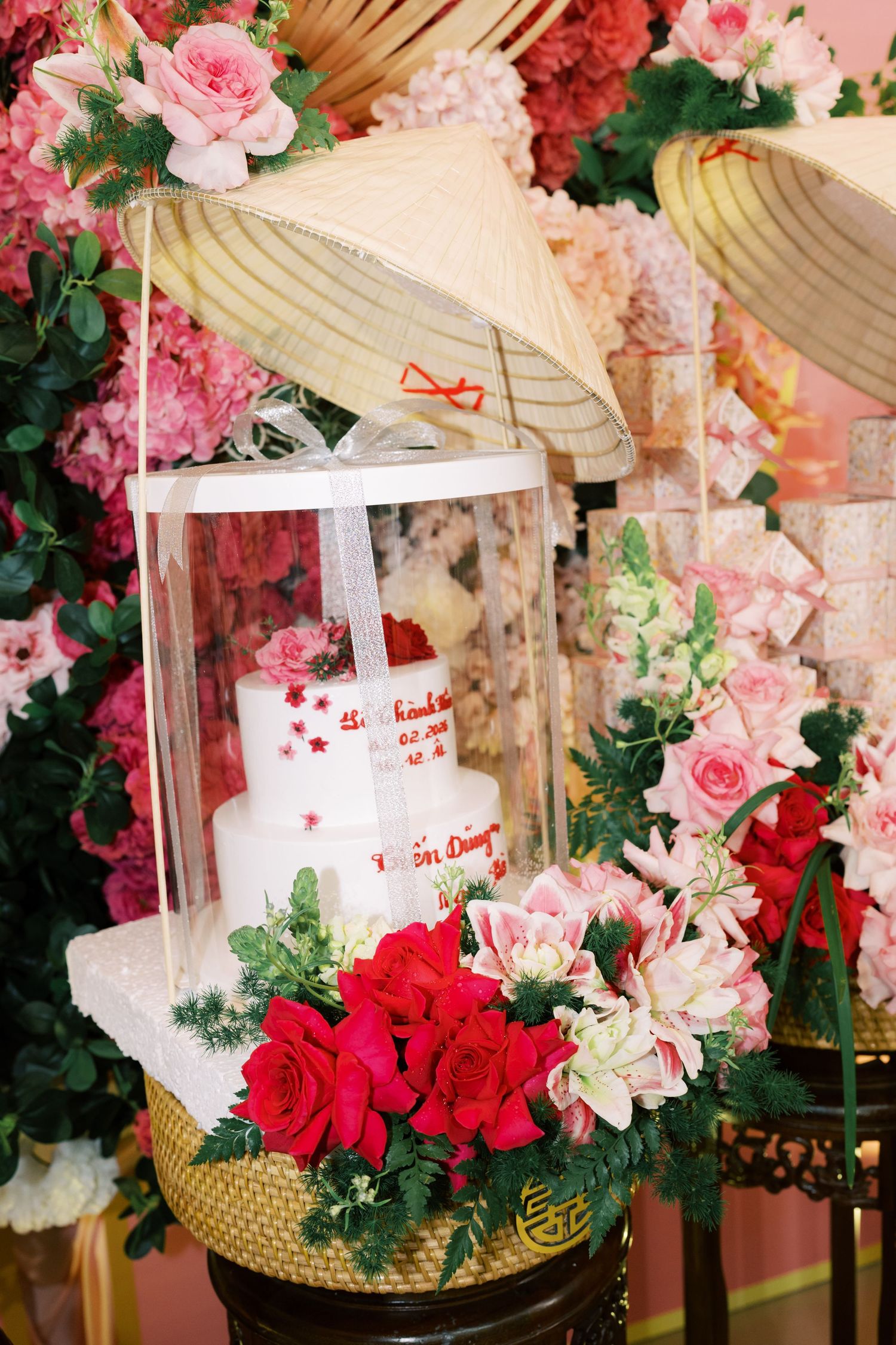 Vietnamese wedding cake displayed in a glass box surrounded by red roses and pink flowers with traditional conical hats.