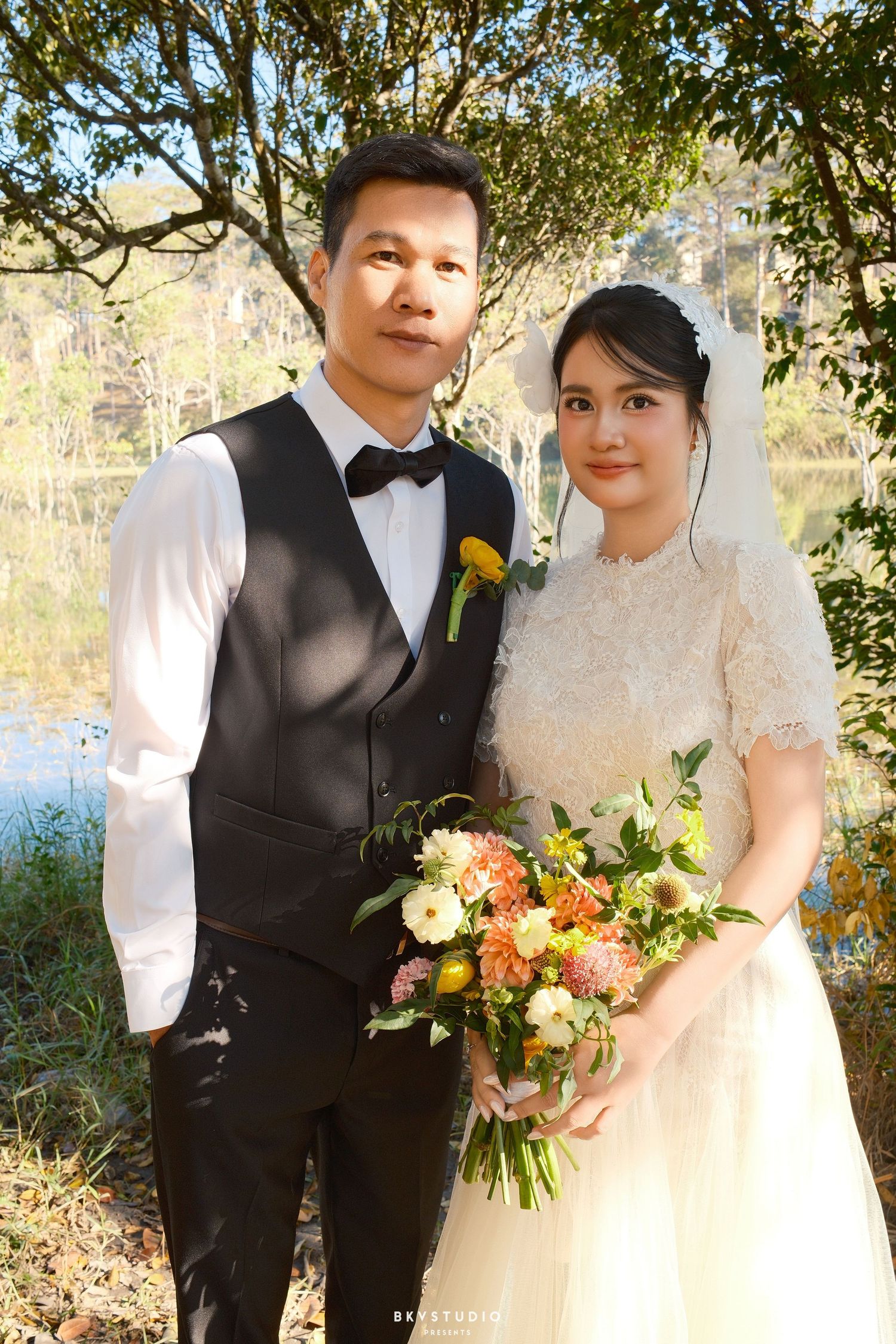 Couple poses in wedding attire with colorful bouquet beside a sunlit lake surrounded by autumn trees.