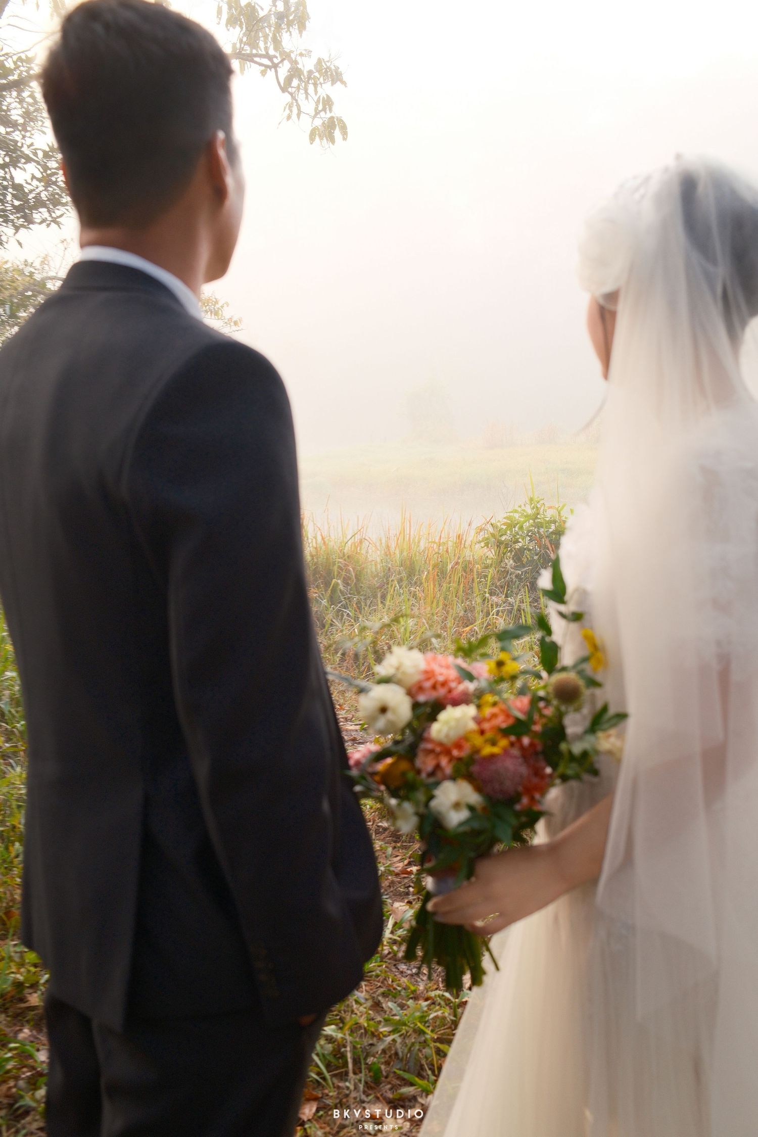 A bride and groom stand together in misty outdoor scenery, bride holding a colorful bouquet and wearing a veil.