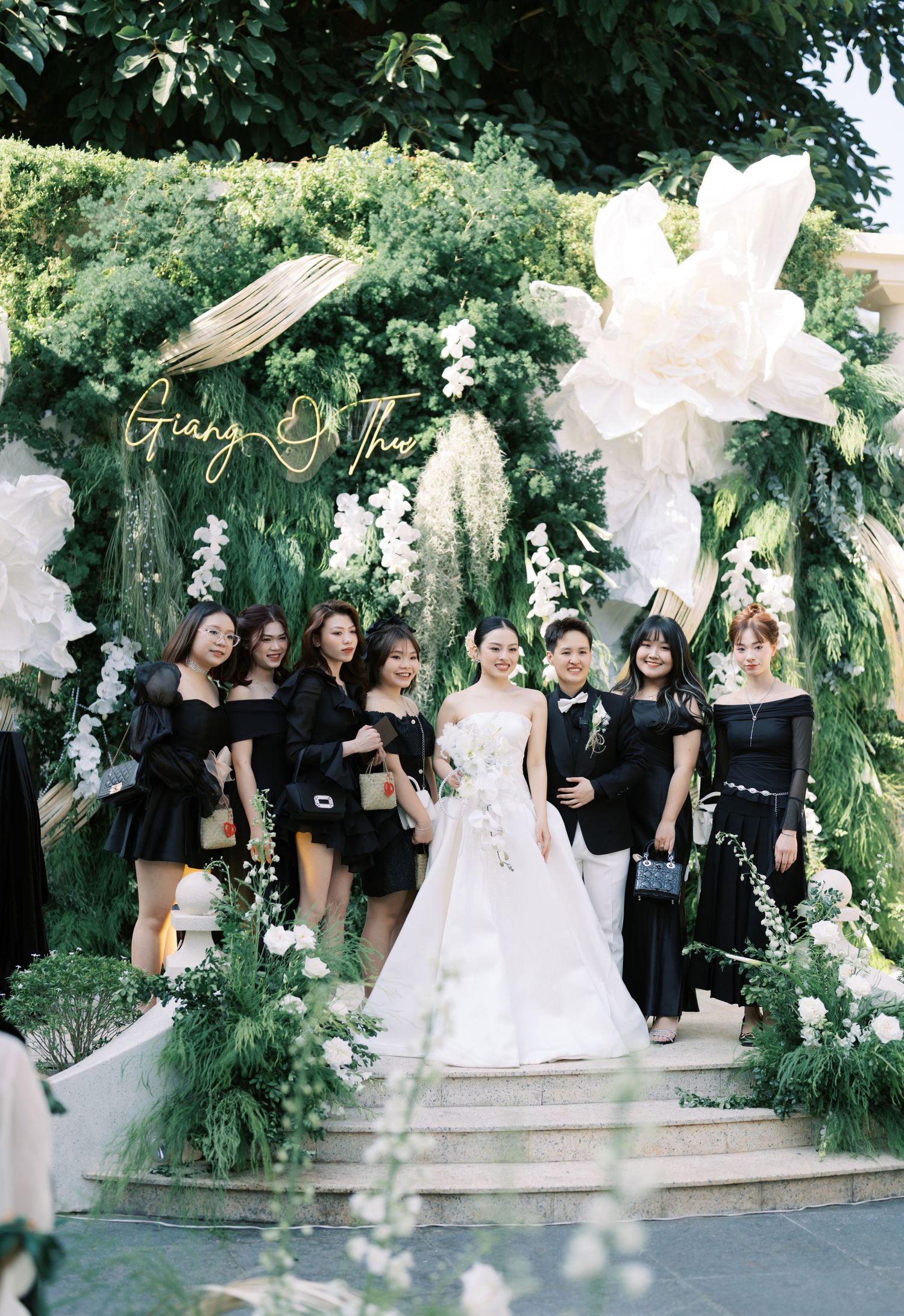Bride in white gown poses with bridesmaids in black dresses at floral wedding arch.