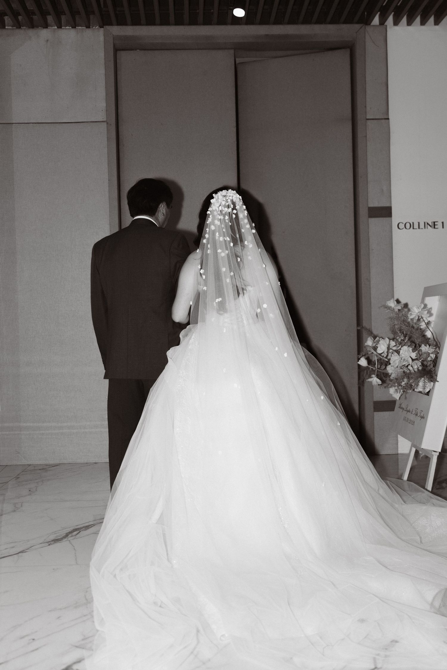 Black and white photo of bride in pearl-adorned veil and ball gown with groom entering wedding venue.