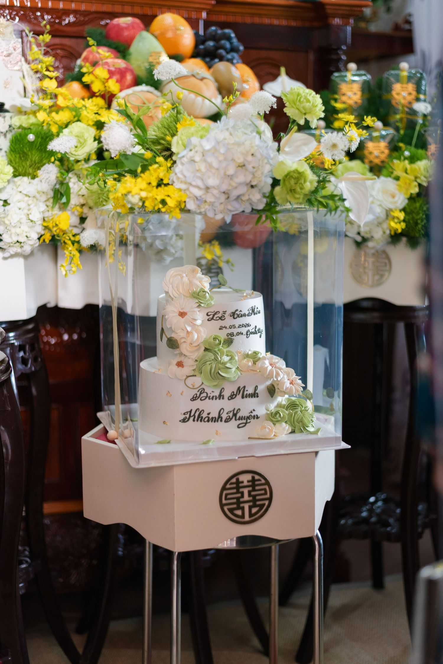 Vietnamese wedding cake displayed in acrylic box with floral arrangements and double happiness symbol.