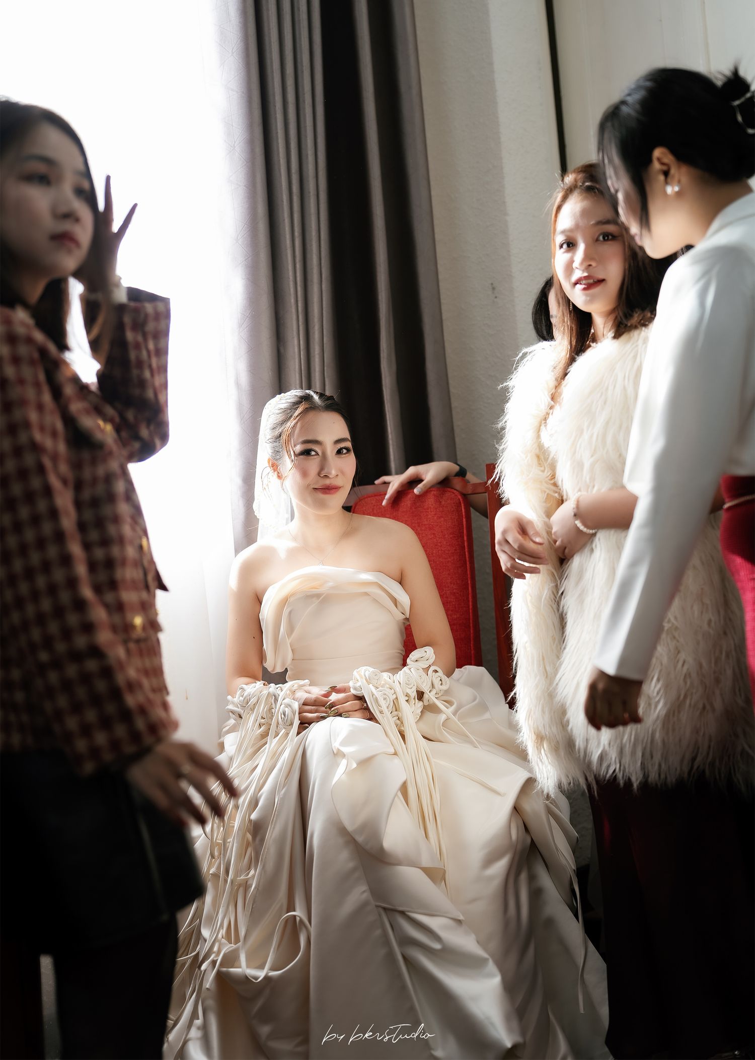 Bride in strapless ivory gown sits in red chair surrounded by bridesmaids during wedding preparations.