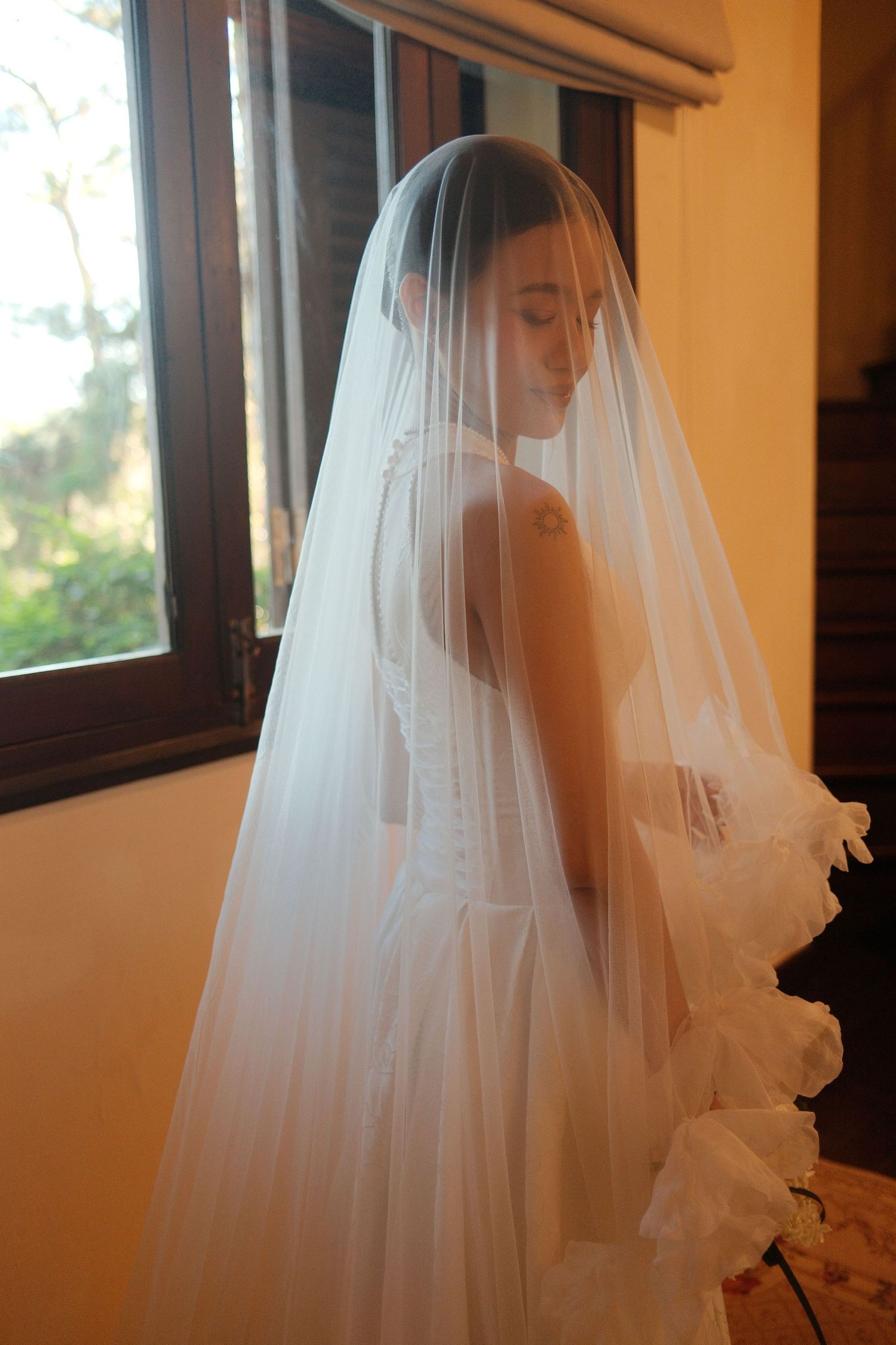 An elegant bride in a white wedding gown and long veil stands gracefully by a sunlit window.