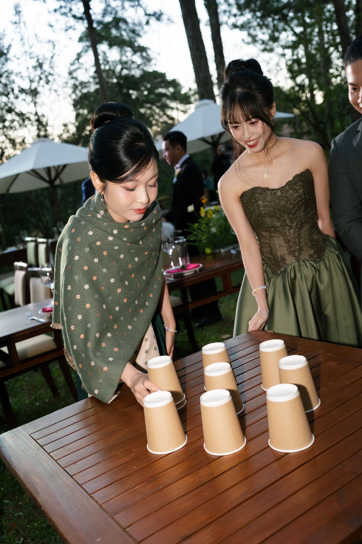 Wedding guests playing cup stacking game at outdoor reception table surrounded by pine trees.