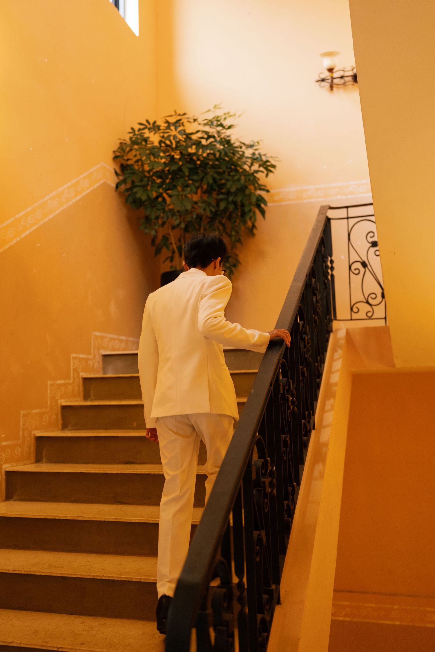 Man in white suit ascending elegant staircase with ornate black railing and green plant against yellow walls.