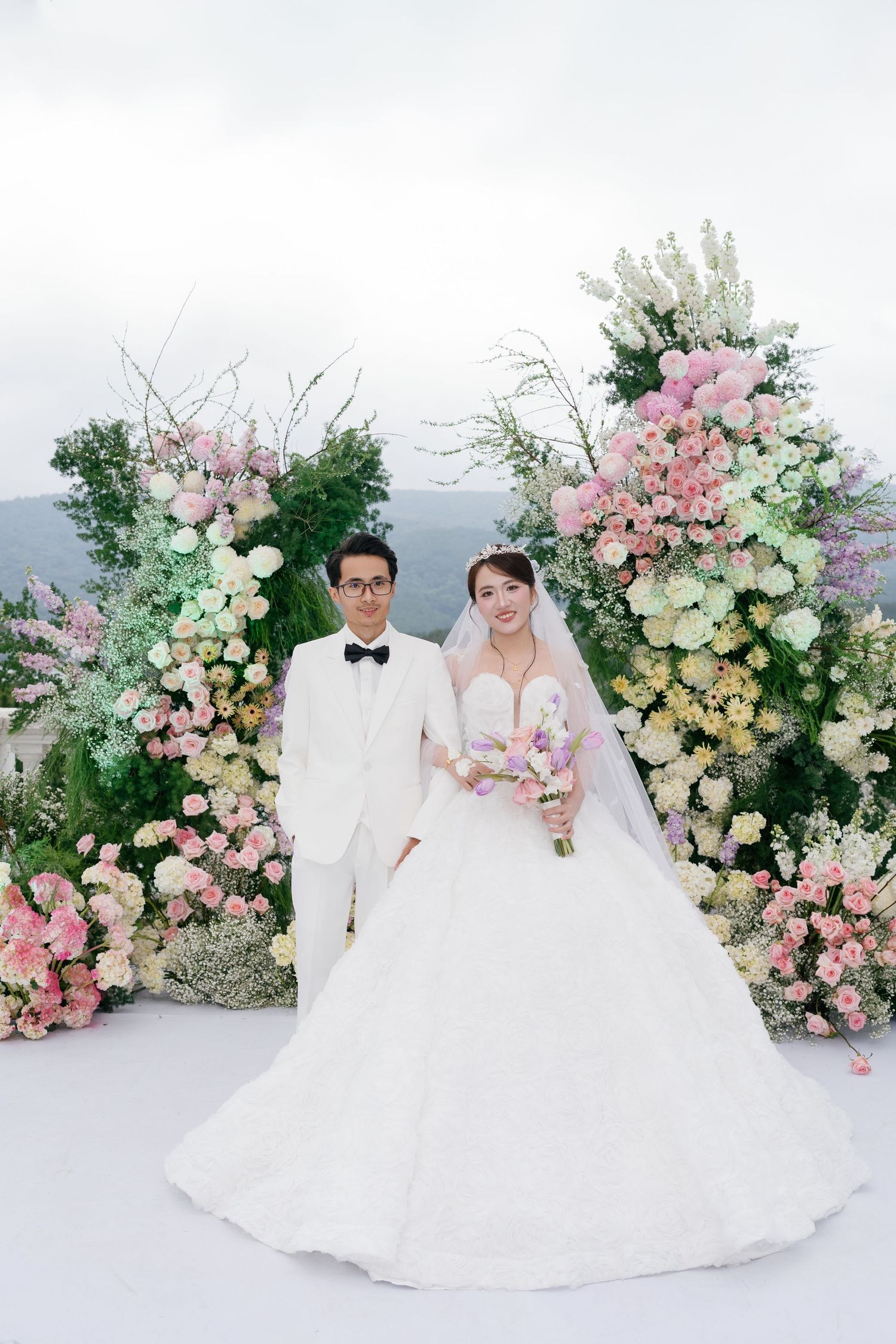 Bride and groom pose before lush floral arch with pink, white, and yellow blooms at an elegant outdoor wedding ceremony.
