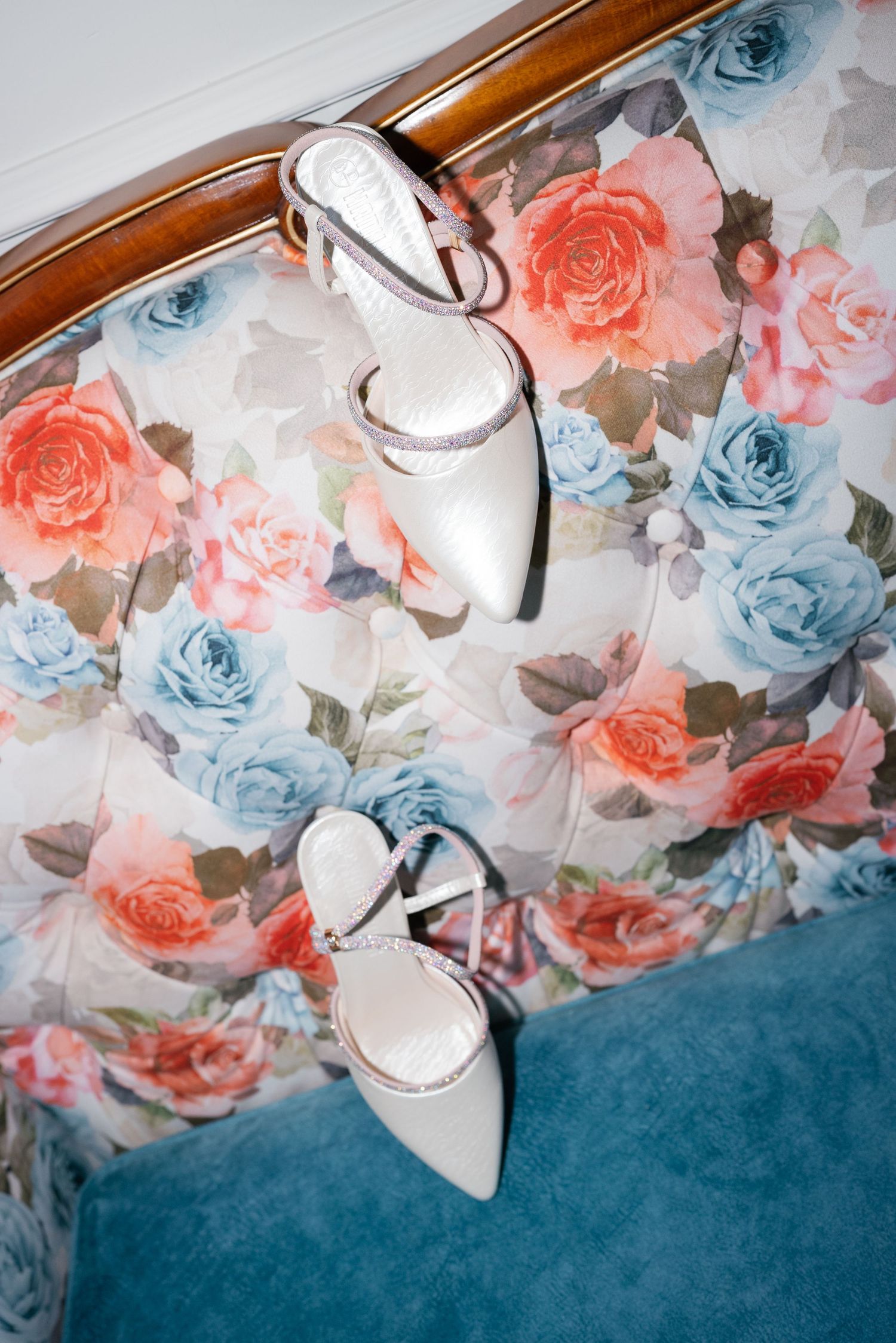 White bridal heels with ankle straps resting on a floral upholstered chair with pink and blue roses.