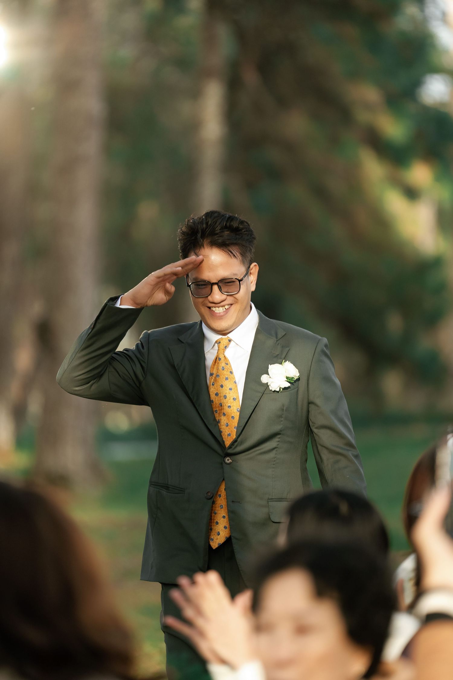 Smiling groom in gray suit and gold tie giving a salute gesture to applauding wedding guests outdoors.