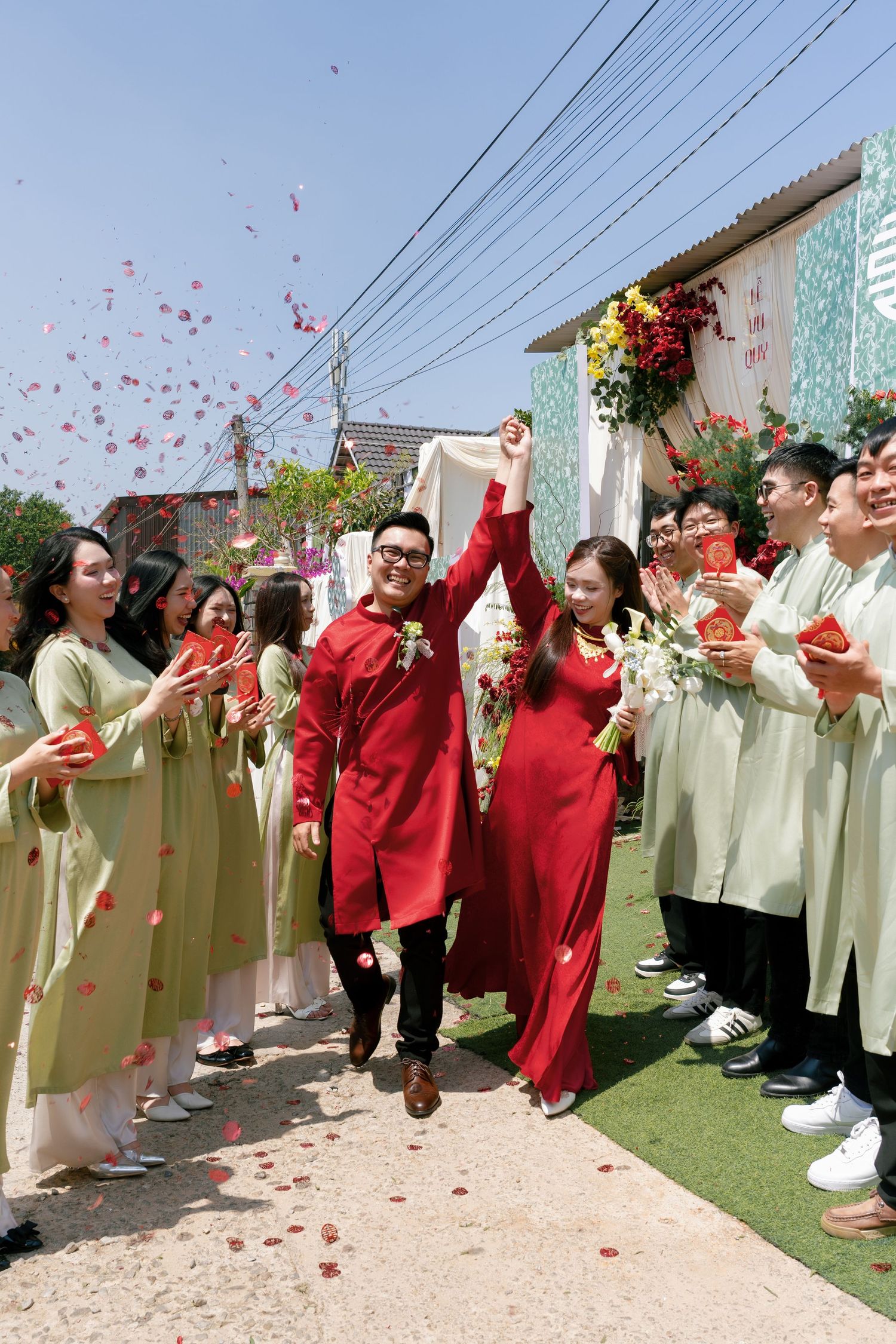 Vietnamese couple in red ao dai celebrate wedding walk with guests in green ao dai tossing flower petals outdoors.