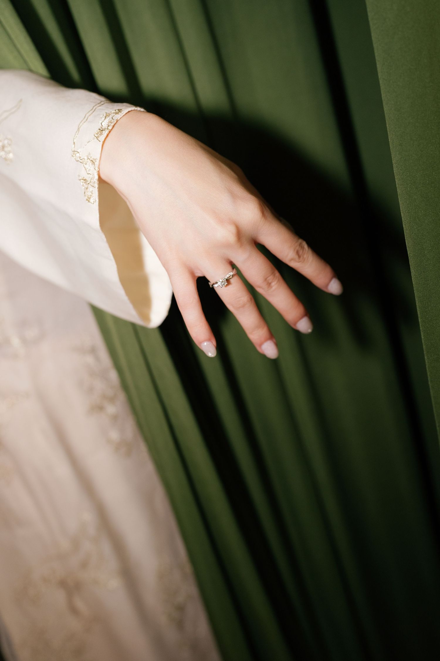 Close-up of a bride's hand wearing a diamond engagement ring and gold bracelet against a green curtain backdrop.