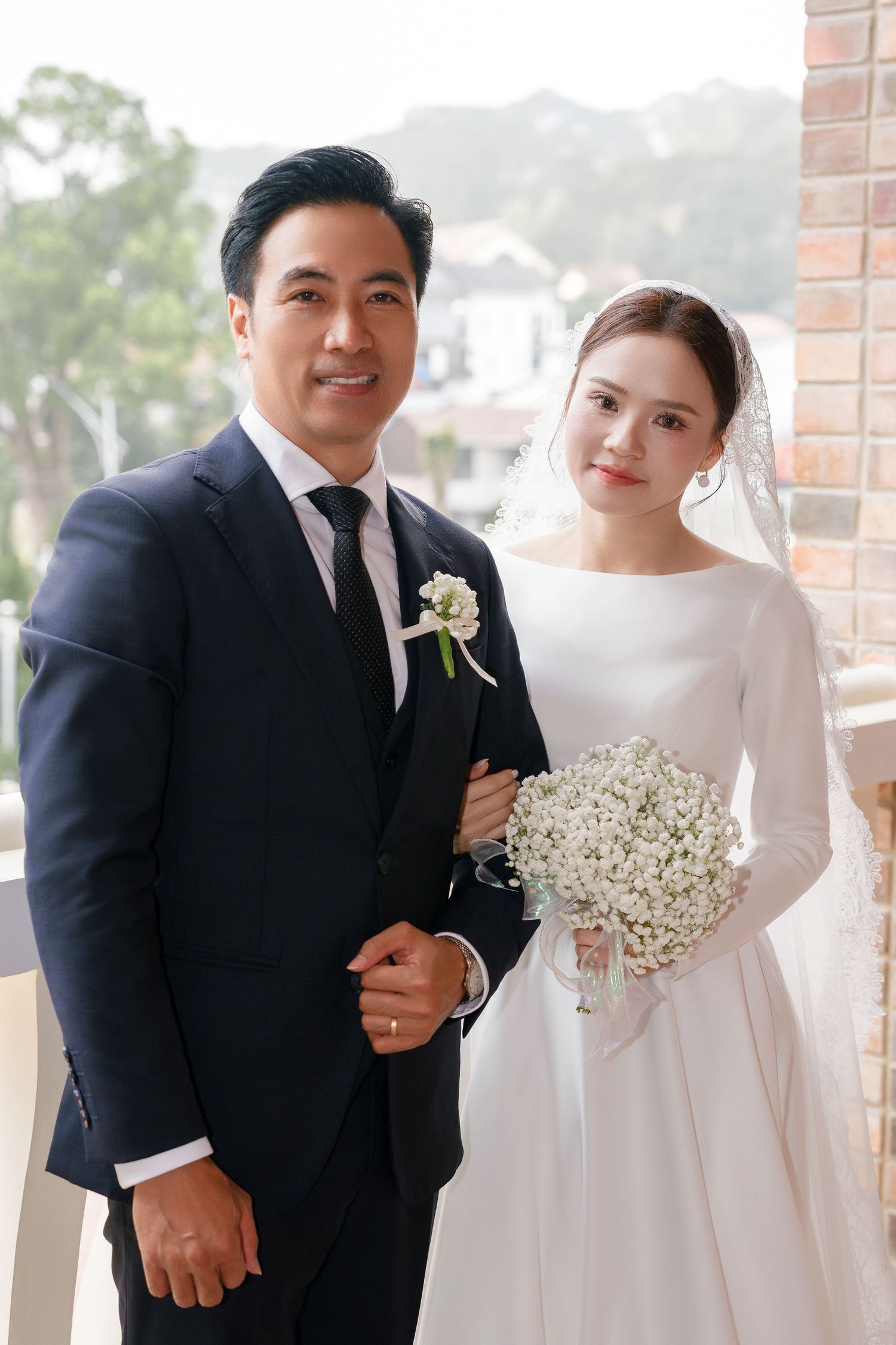Smiling groom in navy suit poses with bride in elegant white dress holding baby's breath bouquet.