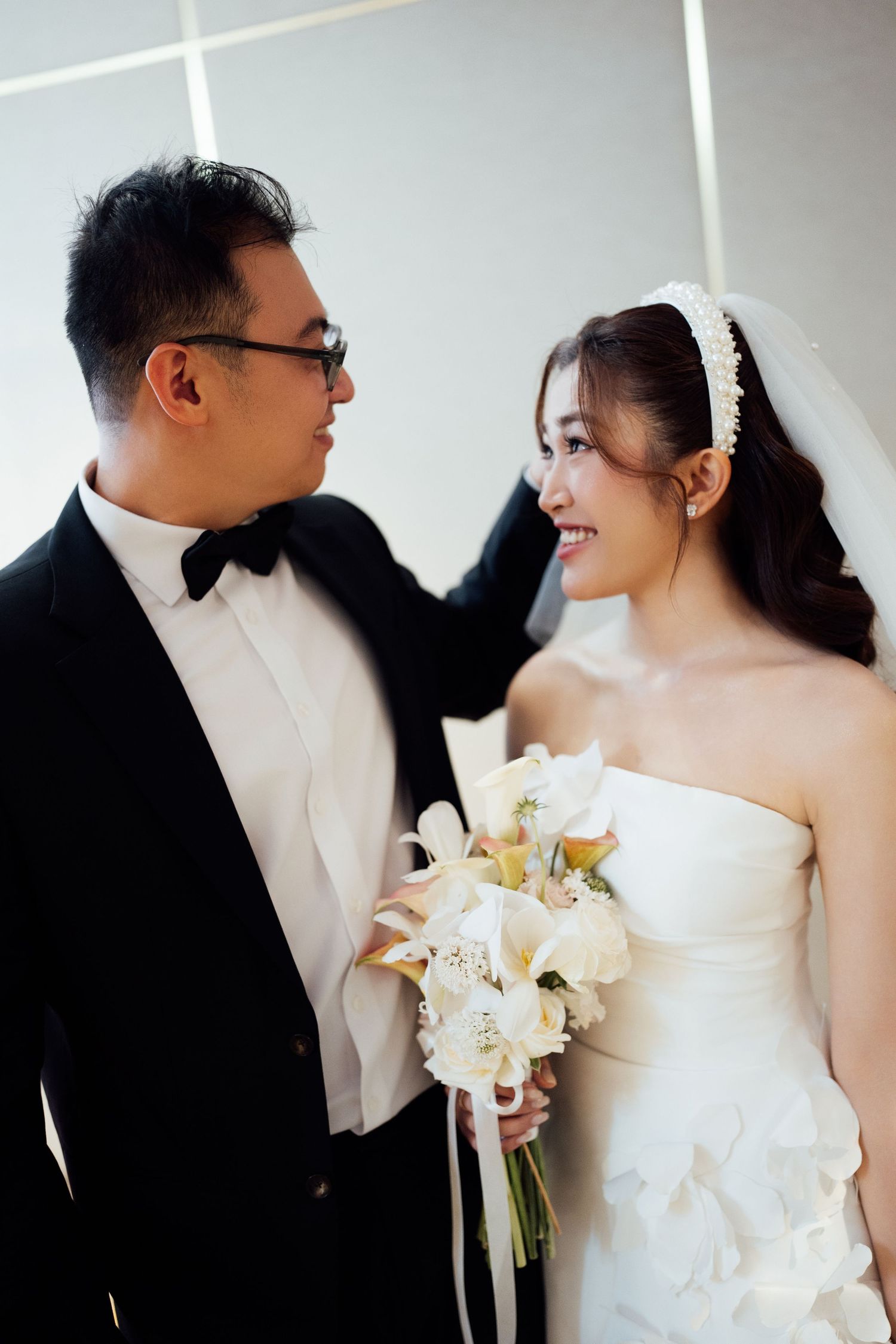 Groom in tuxedo and bride in white gown holding bouquet smiling at each other.