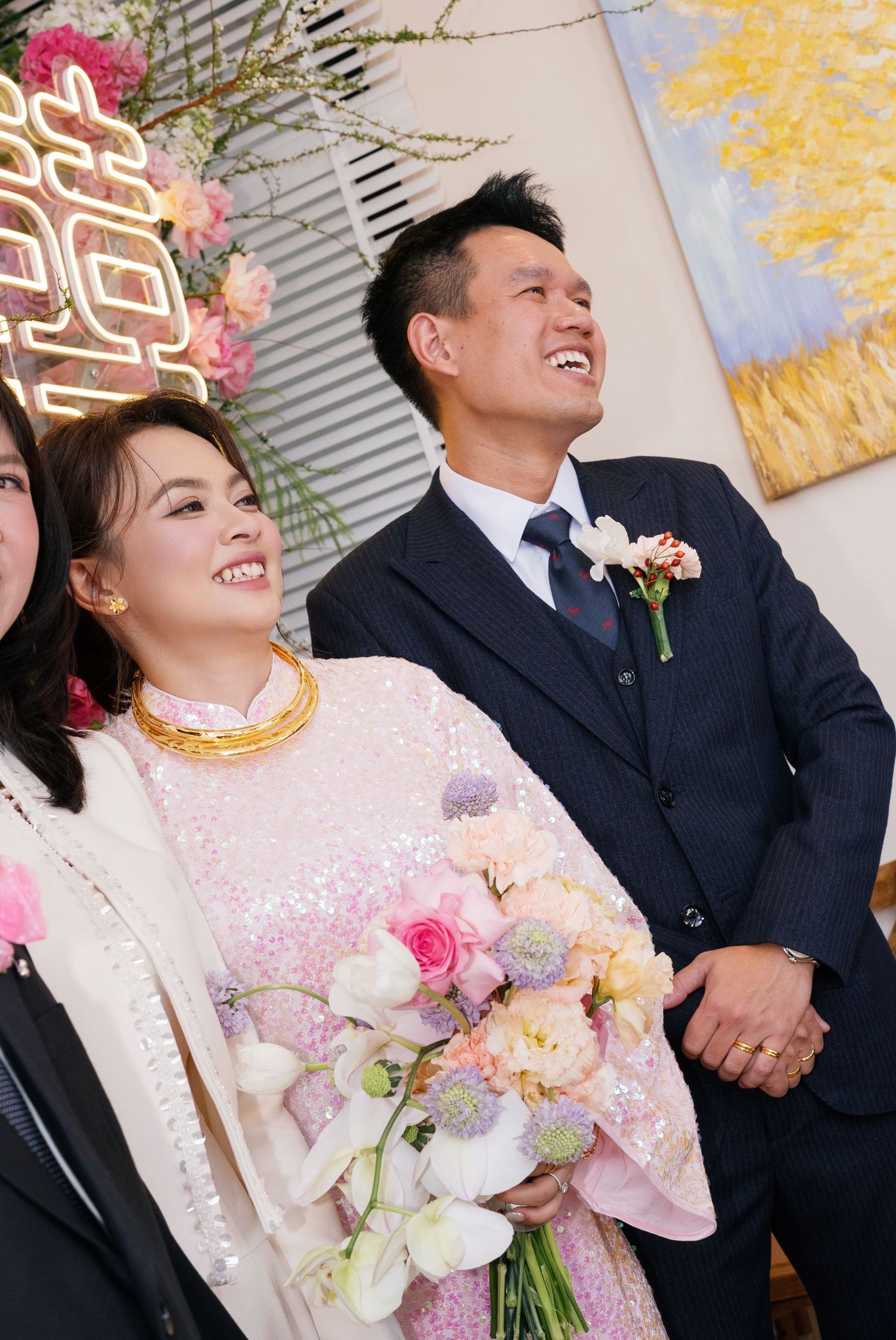 Smiling bride in pink sequin ao dai with gold jewelry holds floral bouquet beside groom in navy suit at Vietnamese wedding.