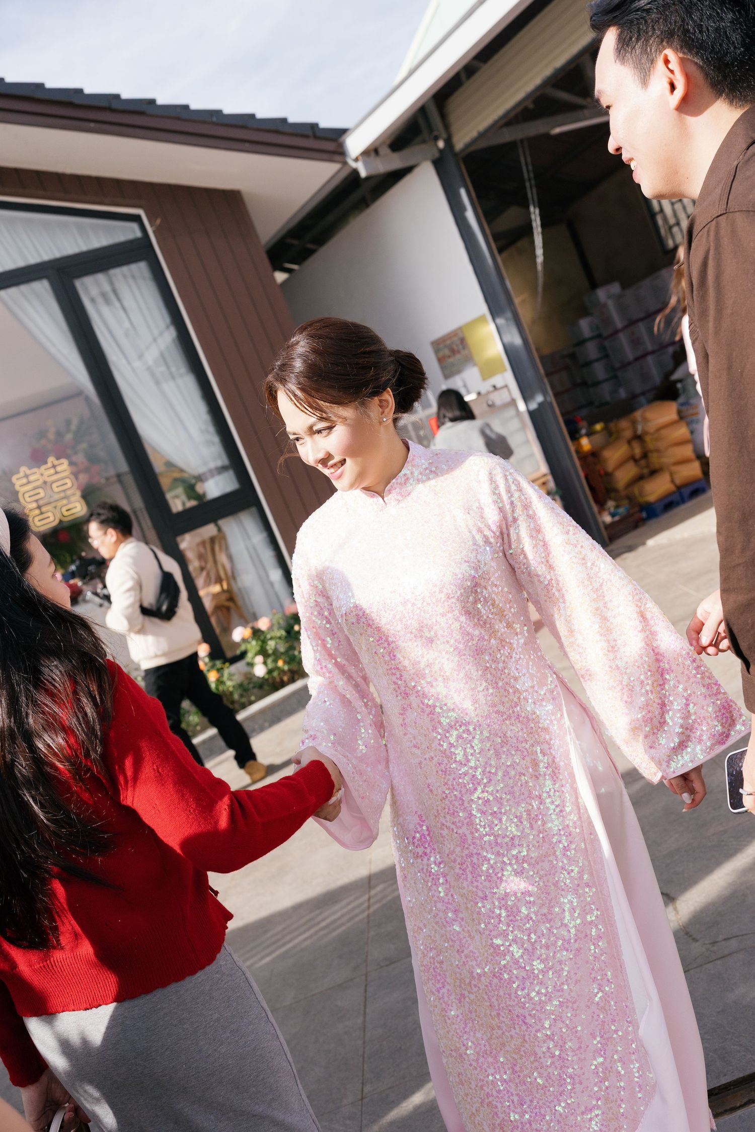 Woman in pink sequined ao dai dress smiling outdoors at a Vietnamese celebration event.