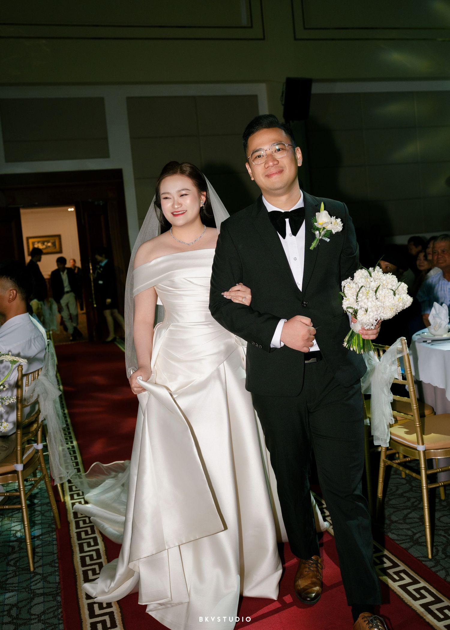 A bride in a white satin gown and groom in a black tuxedo walk arm in arm down the aisle at their wedding ceremony.