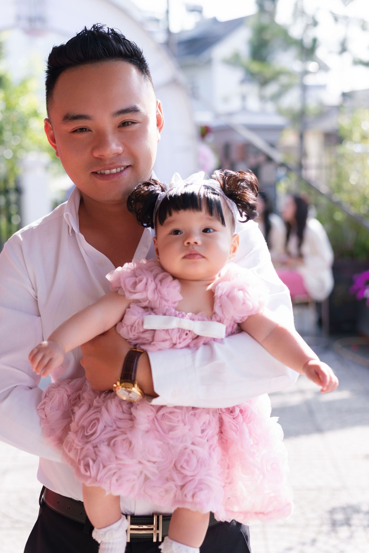 A smiling Asian man holds a cute baby girl in a pink rosette dress with white bow headband outdoors.
