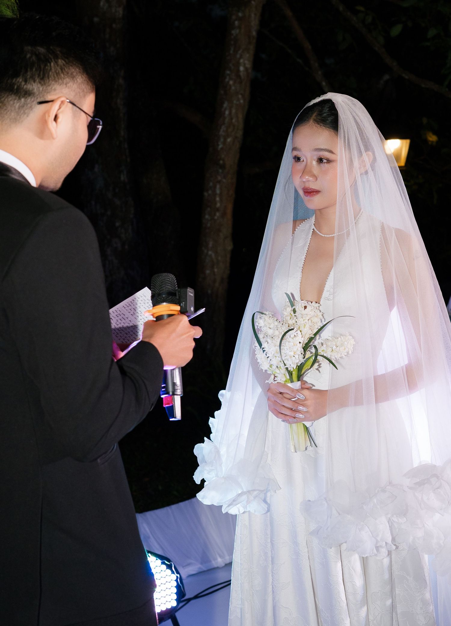 Bride in white veil holding white bouquet listening to officiant during outdoor wedding ceremony at night.