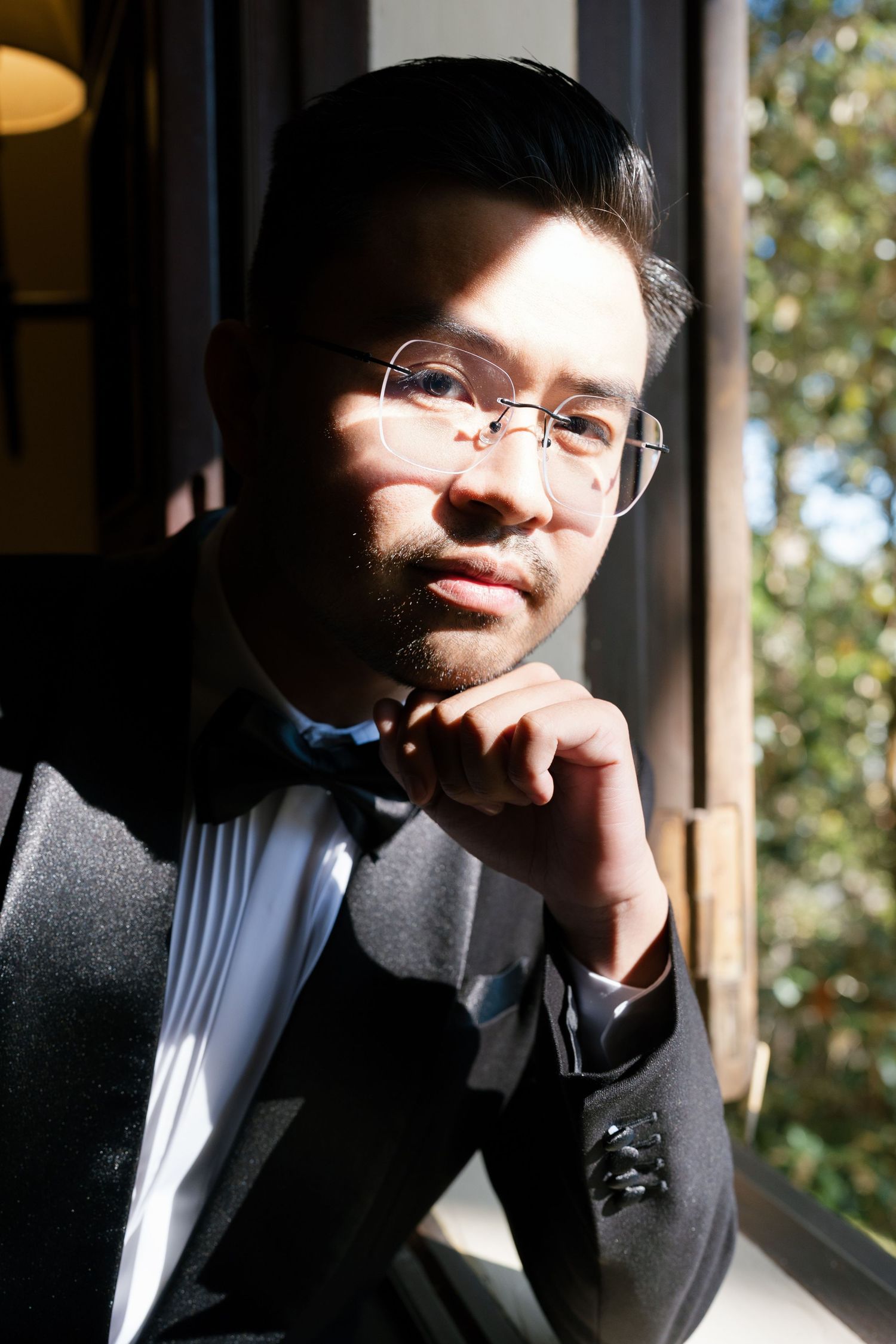 Elegant groom in black tuxedo with bow tie posing thoughtfully by a sunlit window.