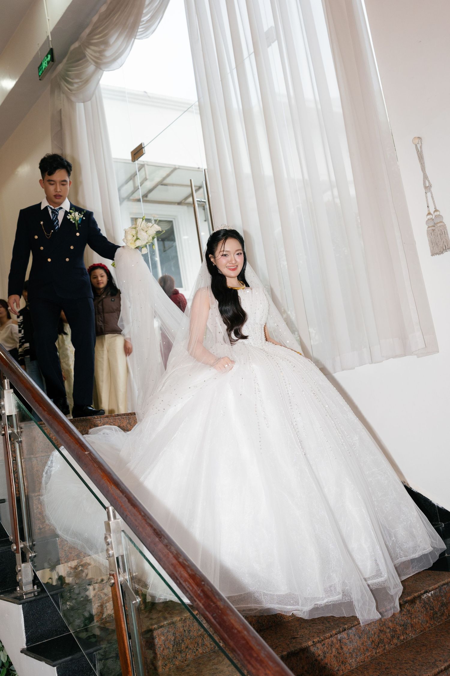 Bride in white ball gown descending staircase with groom in navy suit at elegant wedding venue.