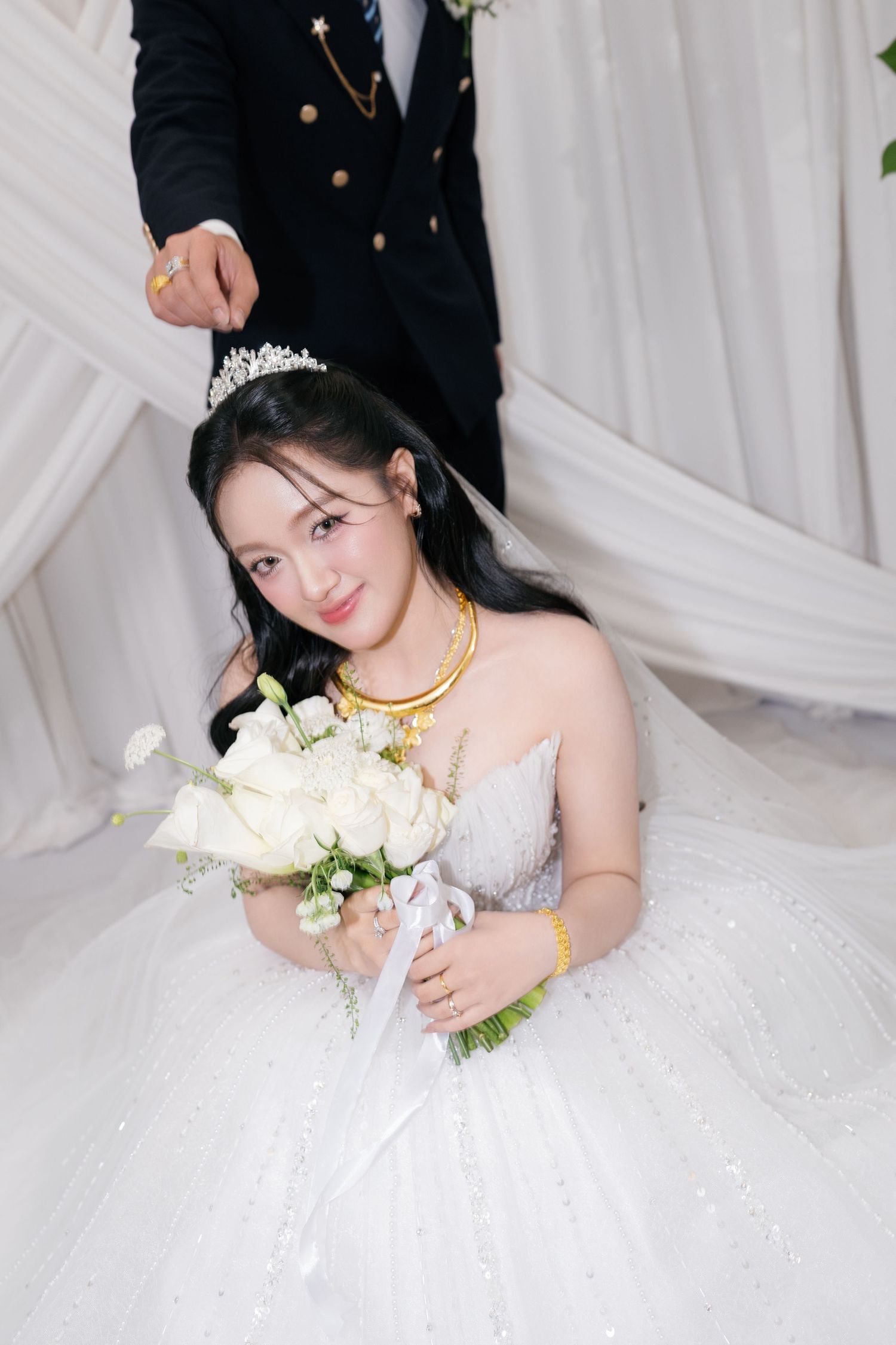 Smiling Vietnamese bride in white ballgown with tiara and gold jewelry holding white rose bouquet.